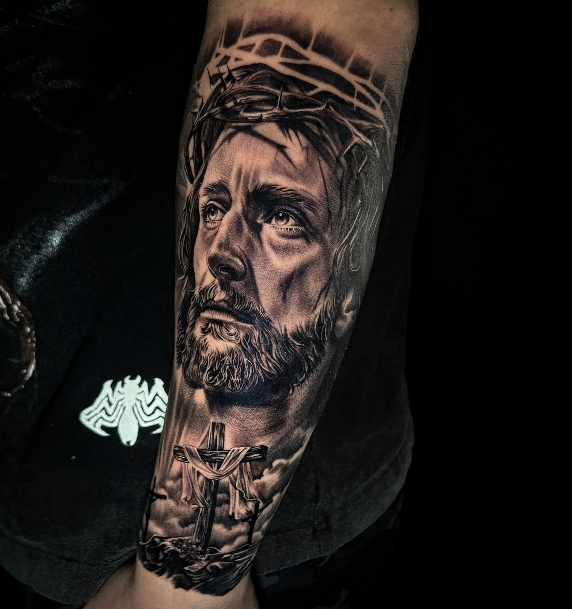 Black and gray realism tattoo of Jesus Christ wearing a crown of thorns with a sorrowful expression, with a wooden cross in the background, on a person's arm.