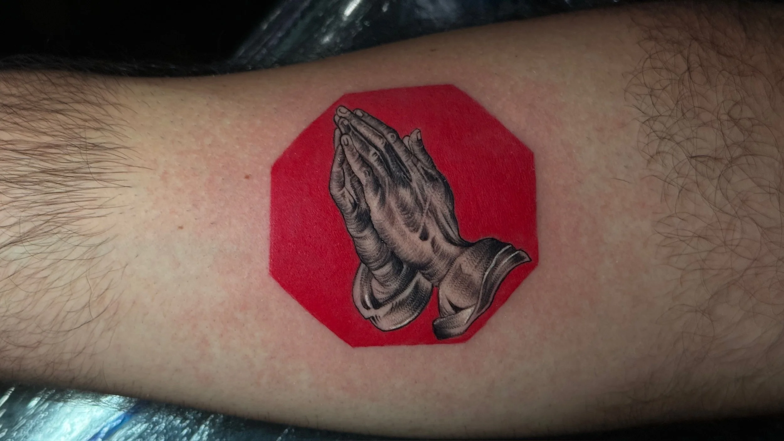 Tattoo of praying hands inside a red octagon shape on a person's arm.