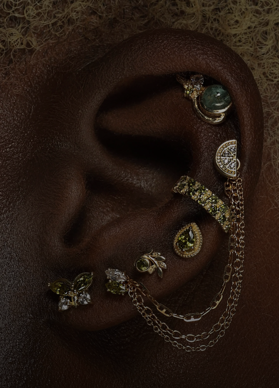 Close-up of a person's ear adorned with multiple gold and silver earrings, jewelry chains, and other decorative jewelry pieces.