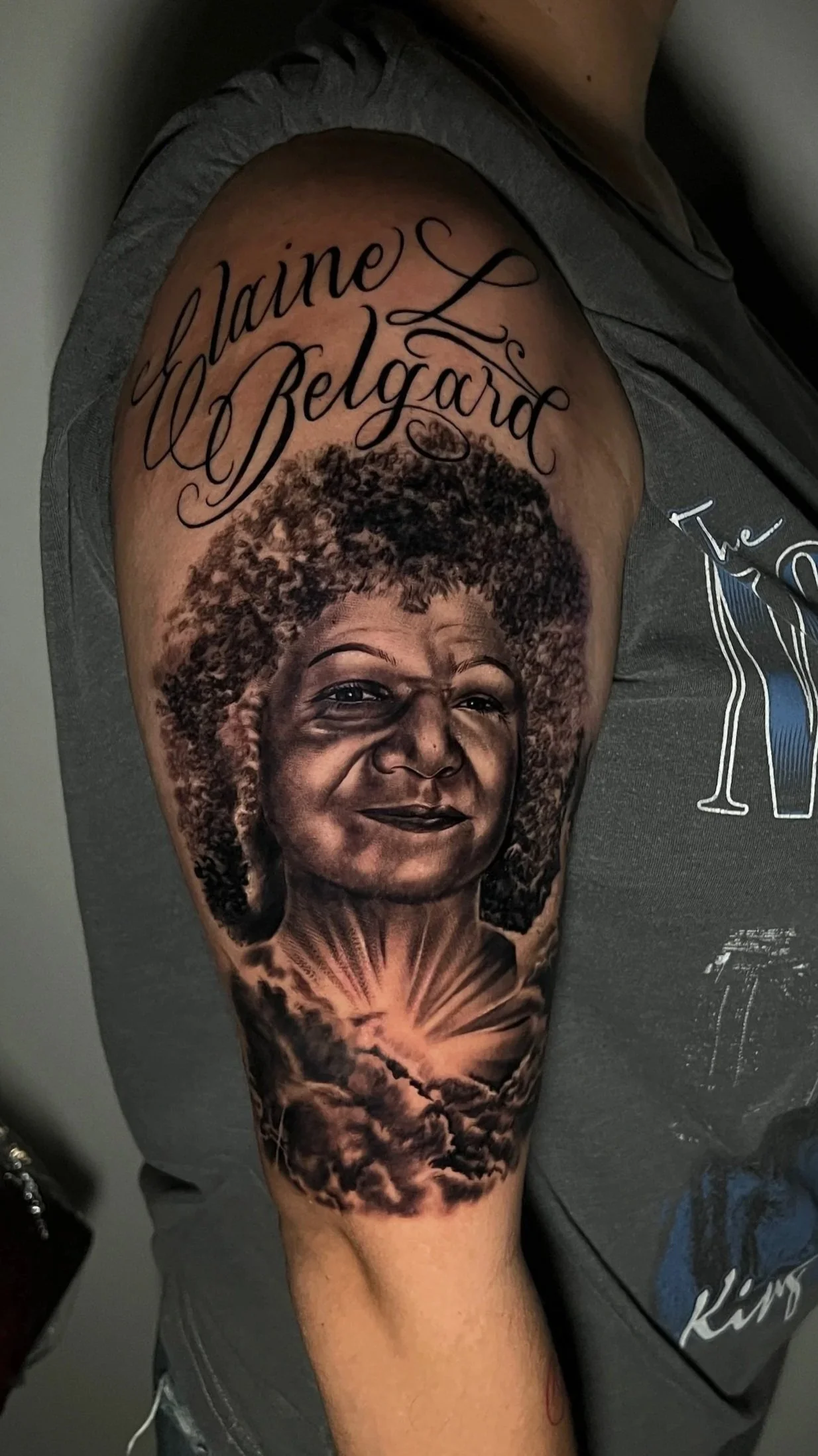 Tattoo of a woman's face with curly hair, smiling, on upper arm. Above the face, text reads 'Elaine L Belgard' in cursive. Below, a realistic depiction of a sunrise or sunset with clouds.