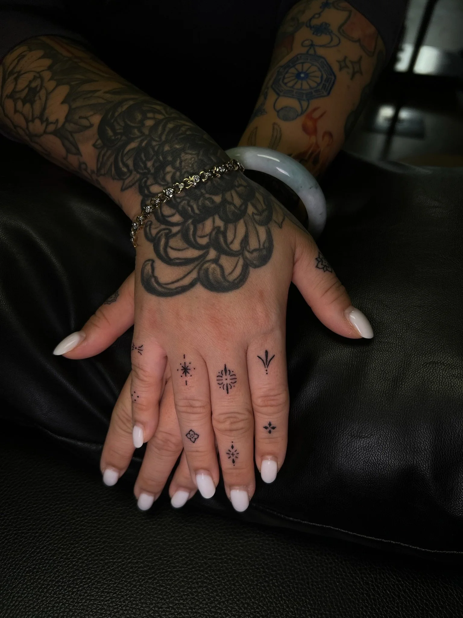 Close-up of a person's hand with tattoos, white French manicured nails, rings, and decorative finger tattoos, resting on black leather surface. The person's wrist has a bracelet, and the background shows more tattoos on the arm.