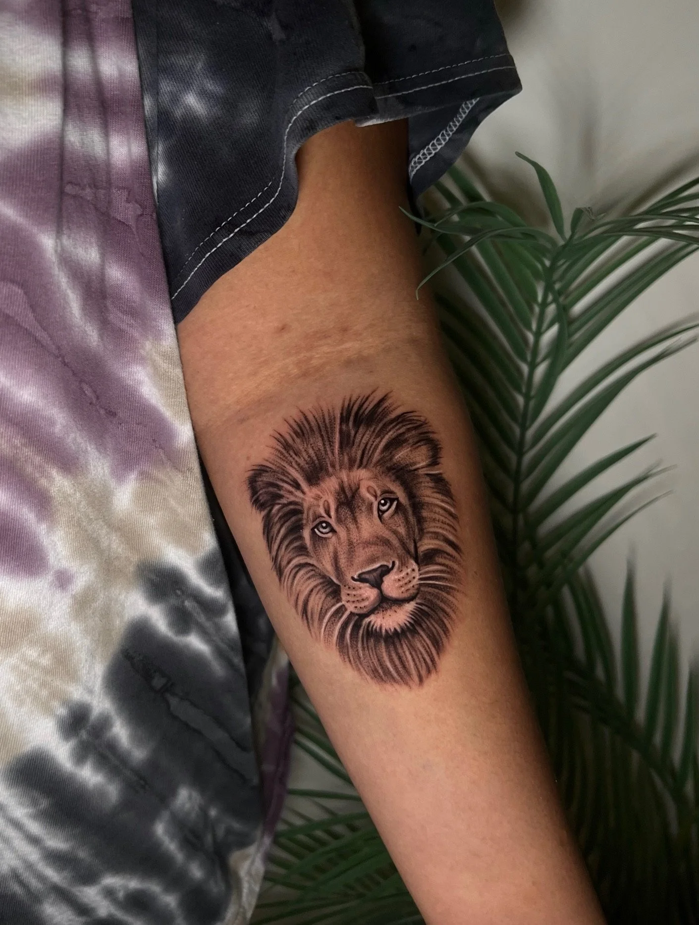 Tattoo of a lion's face on a person's arm, with the person sitting beside some green leaves.