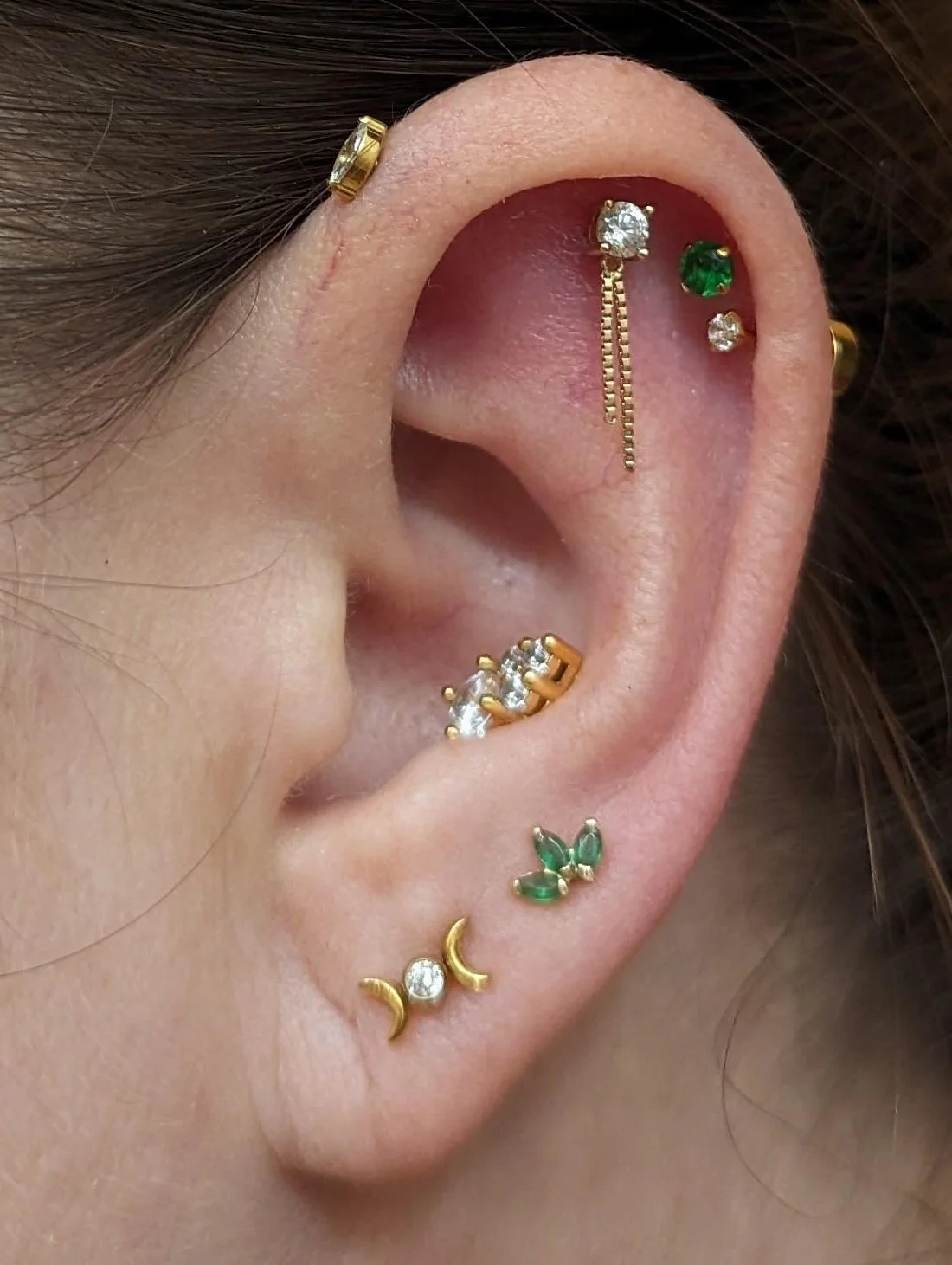 Close-up of a person's ear adorned with multiple gold earrings, including a chain earring with a clear gemstone, a green gemstone stud, a small clear gemstone stud, a curved earring with three clear gemstones, green leaf-shaped earrings, and a crescent moon and gemstone earring.