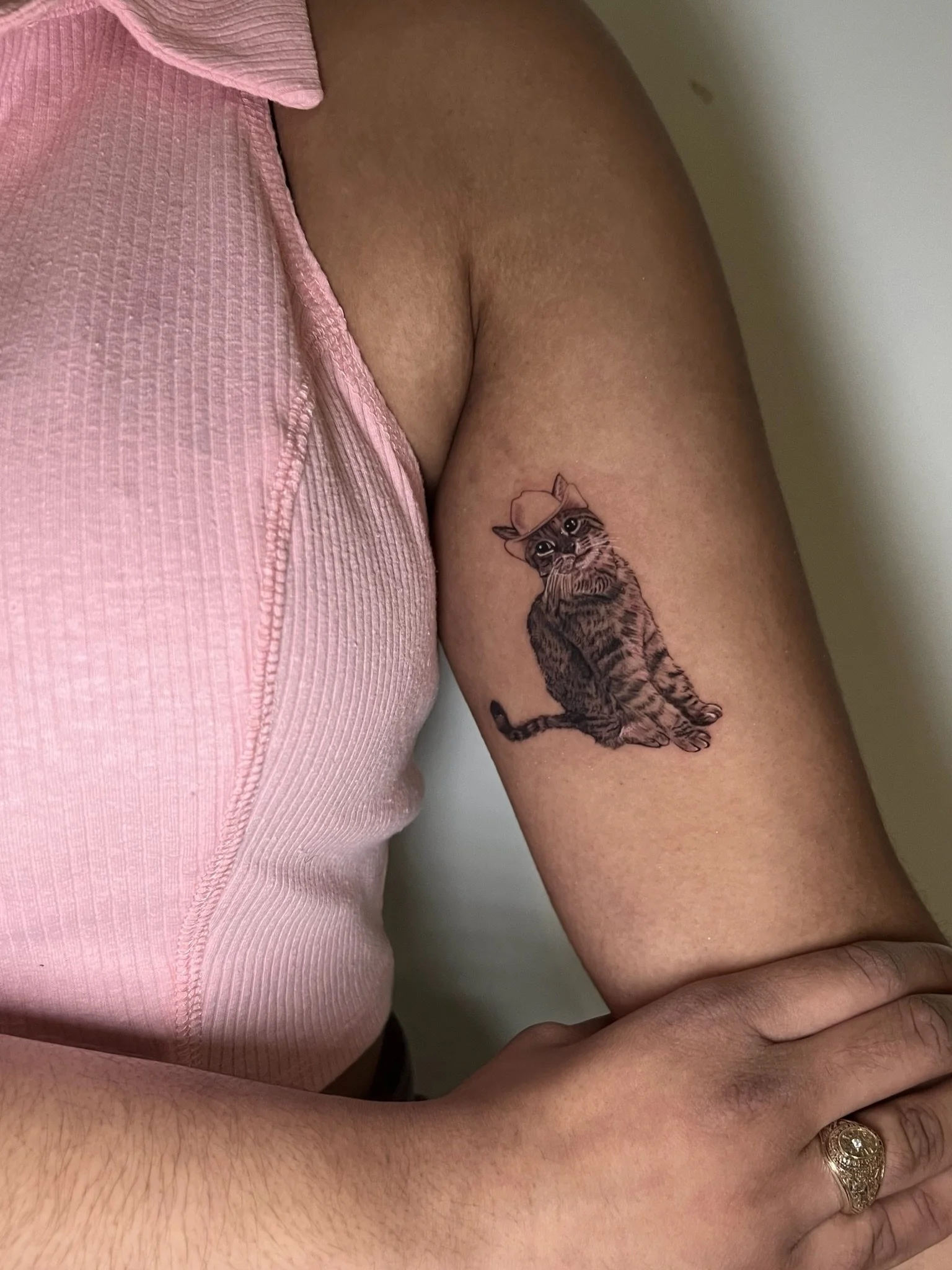 A person's upper arm with a tattoo of a kitten wearing a hat and big eyes.