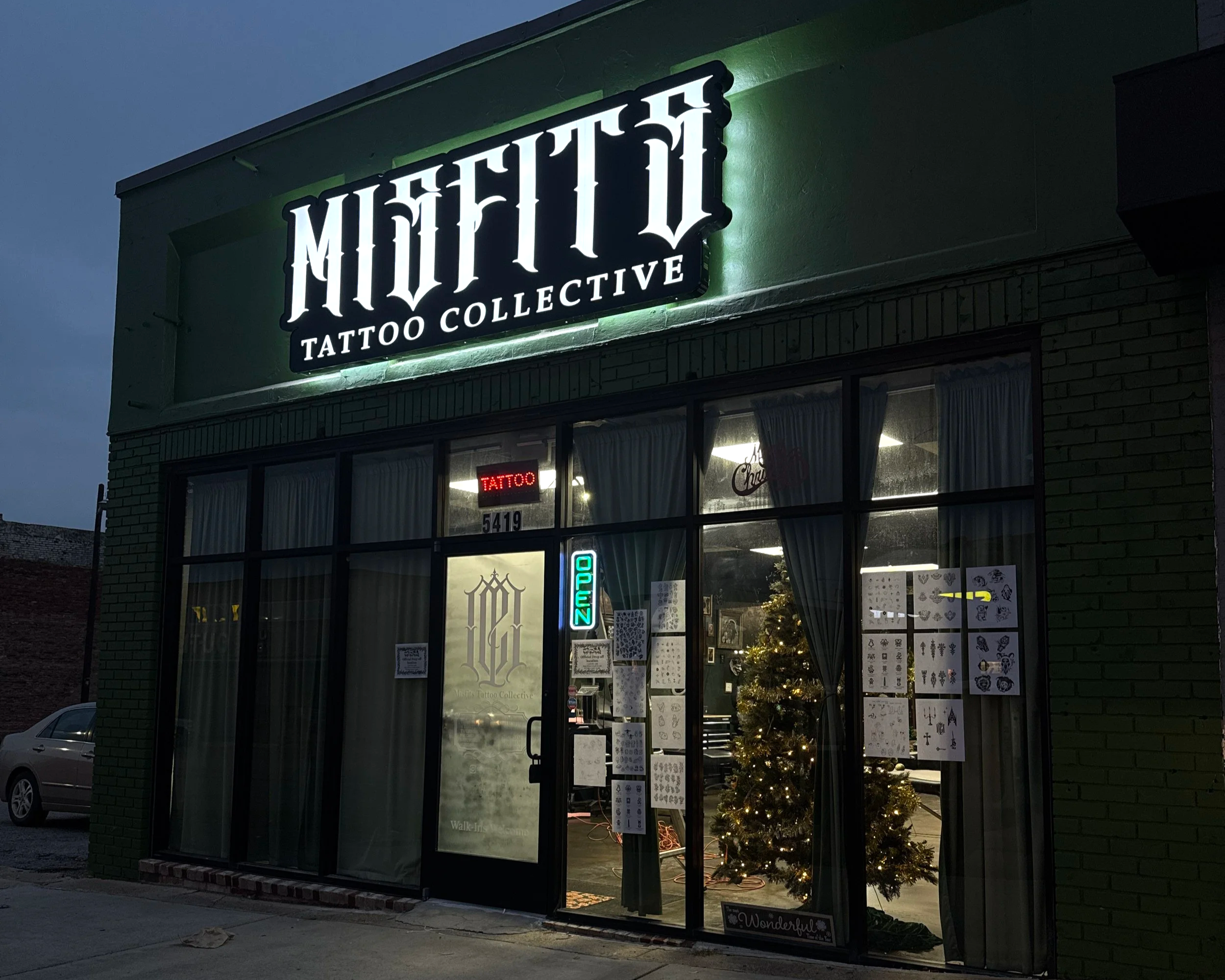Nighttime view of a tattoo shop called 'Misfits Tattoo Collective' with a lit sign, decorated Christmas tree inside, and tattoo designs posted on the window.