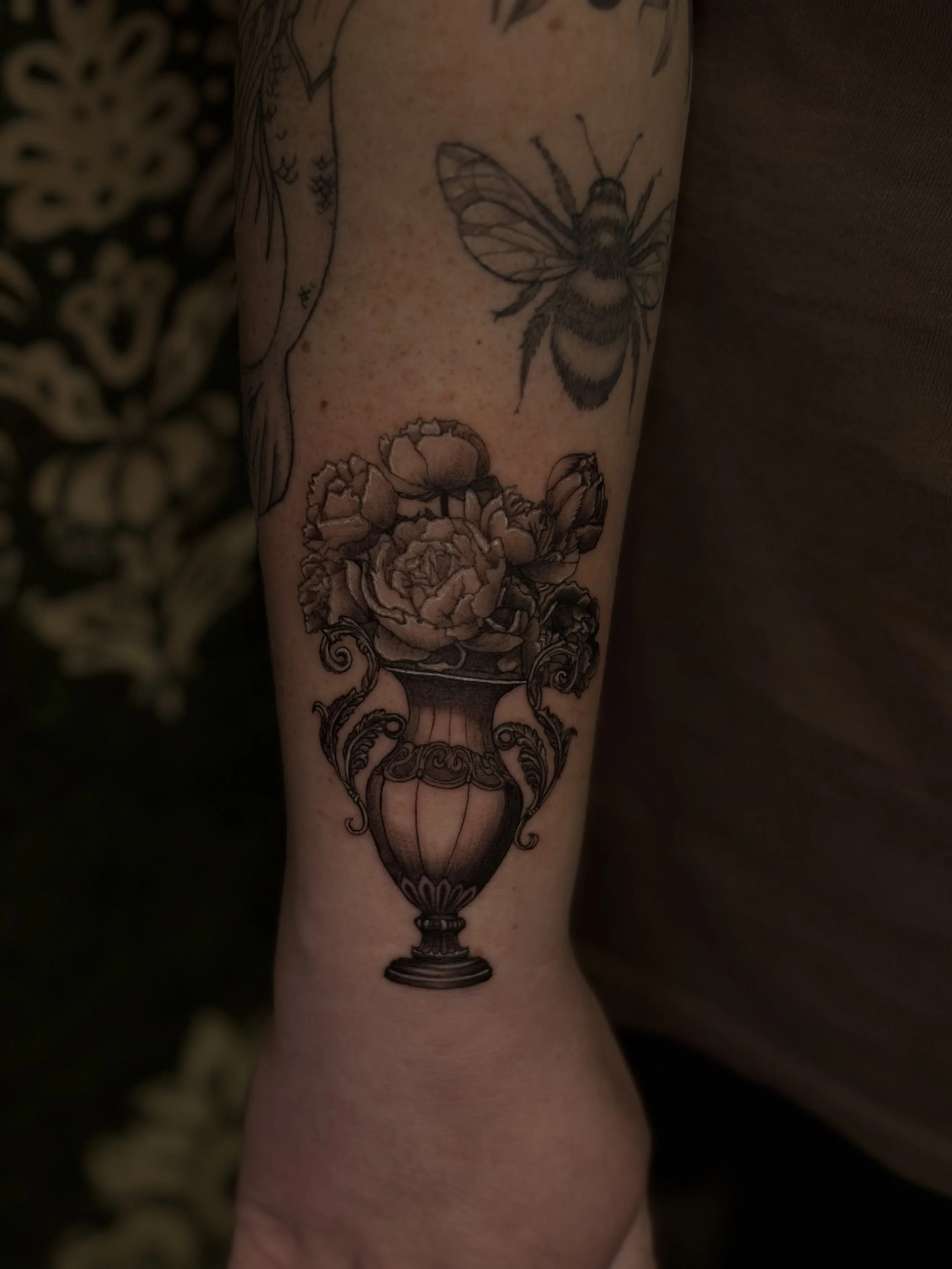 Tattoo of a bee flying above a vase of flowers on a person's arm.