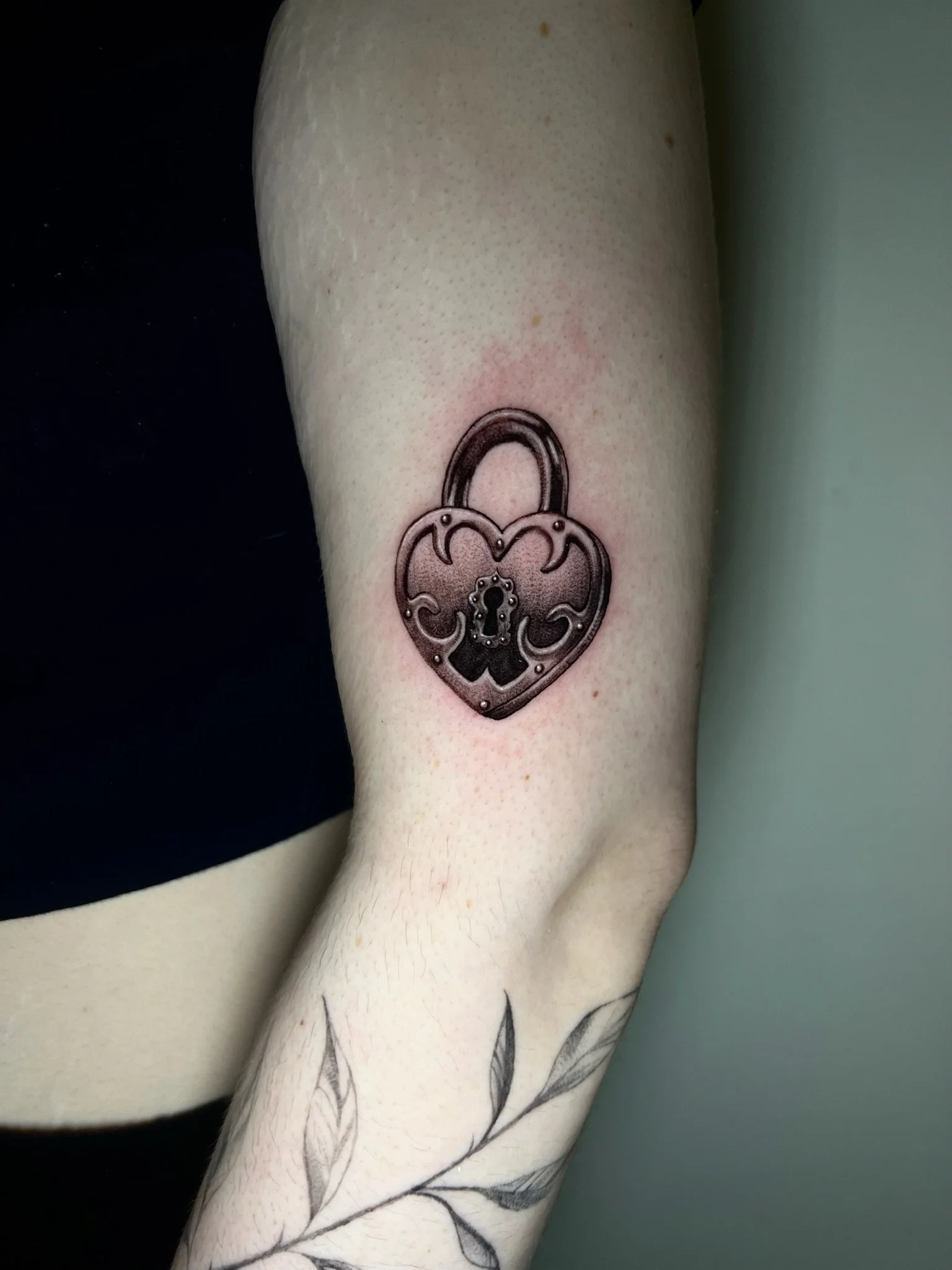 Tattoo of a heart-shaped padlock on upper arm with floral design and skull in the center.