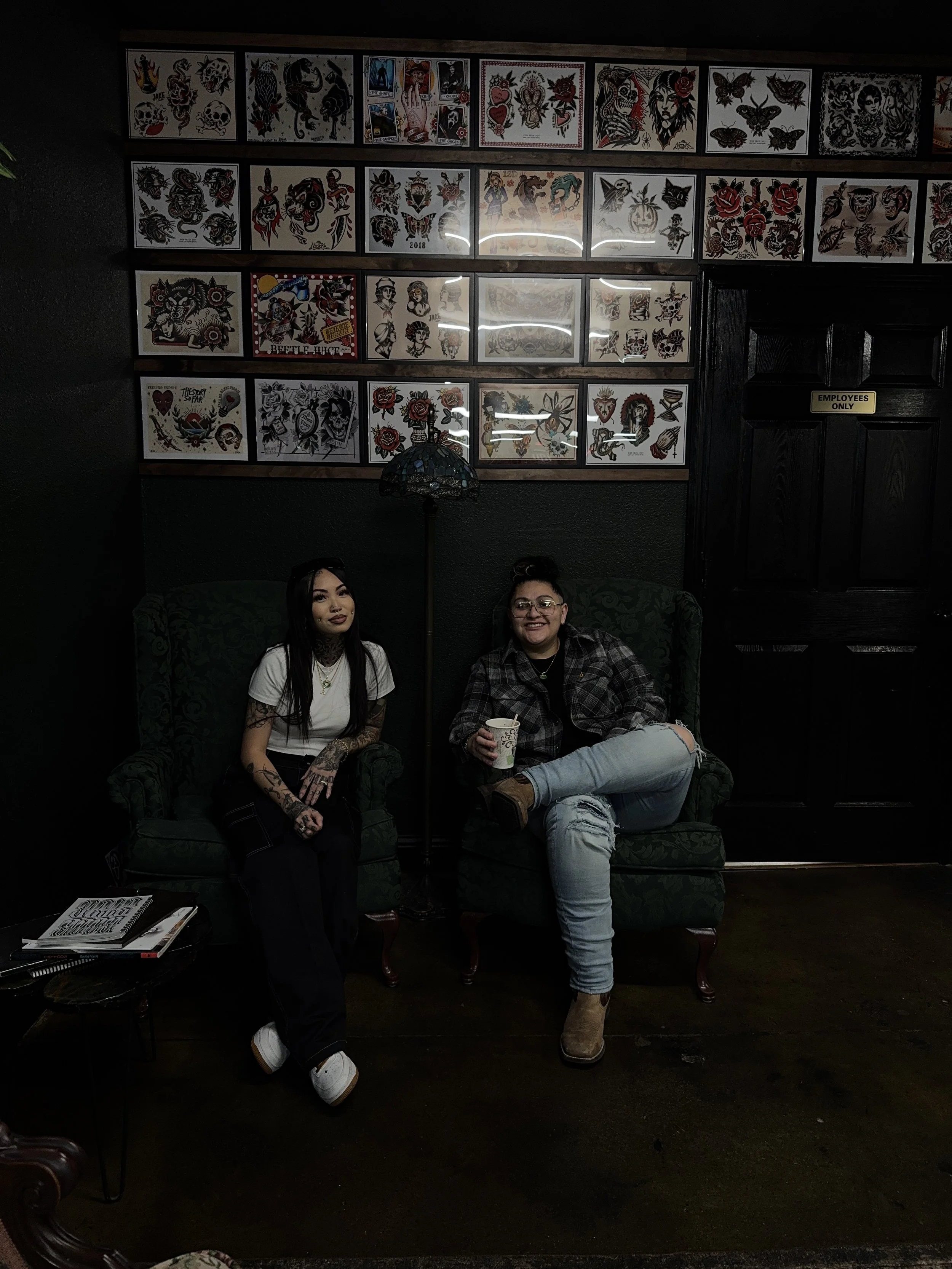 Two women sitting on green patterned armchairs in a dimly lit room with tattoo flash art on the wall behind them. One woman has long dark hair and tattoos on her arms, dressed in a white t-shirt and black pants. The other woman has glasses, dark hair tied up, wearing a plaid shirt and ripped jeans, holding a cup. A small table with notebooks and pens is beside them, and a door labeled 'Employees Only' is in the background.