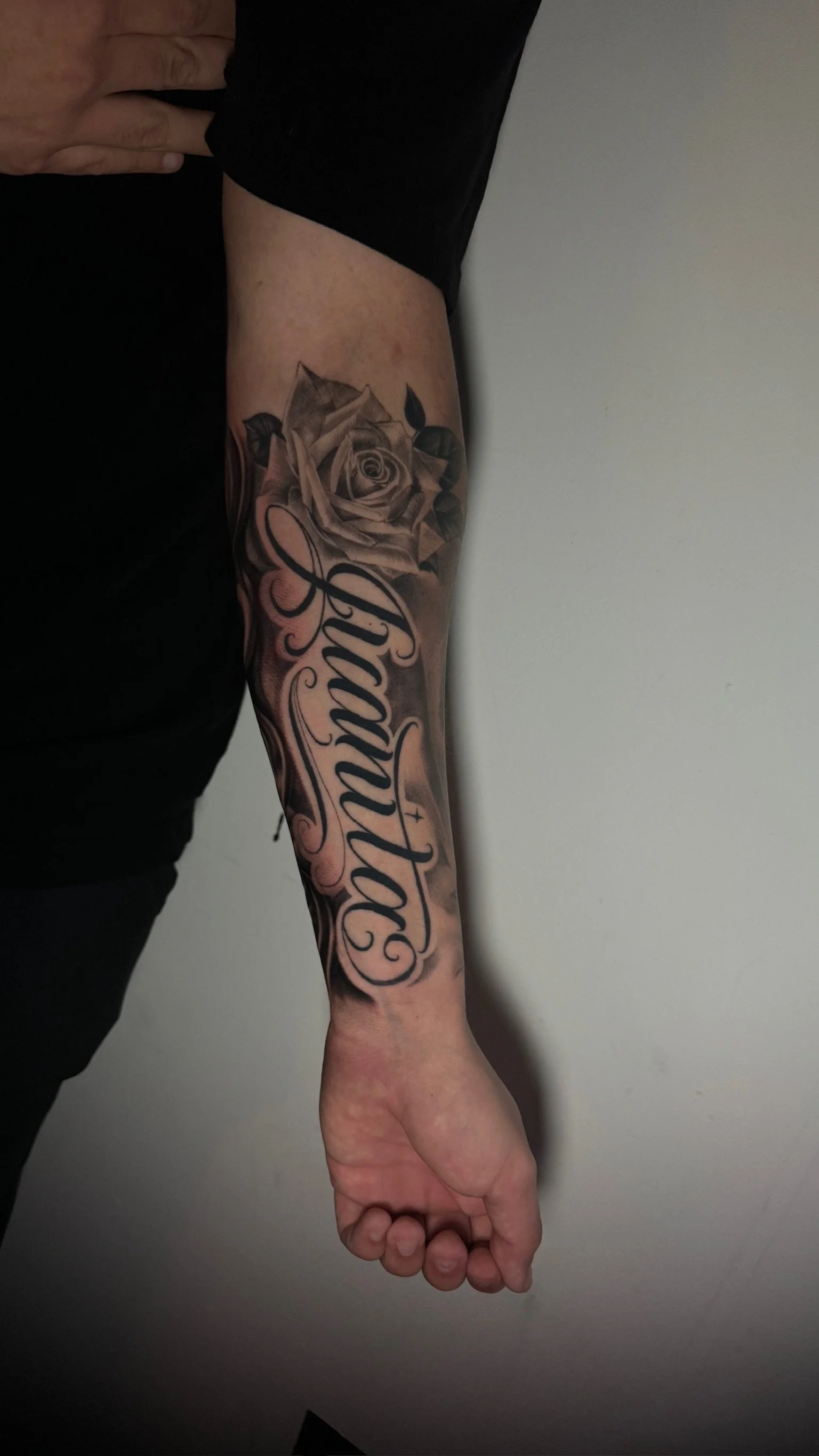 Tattoo on person's forearm featuring a rose and cursive text.