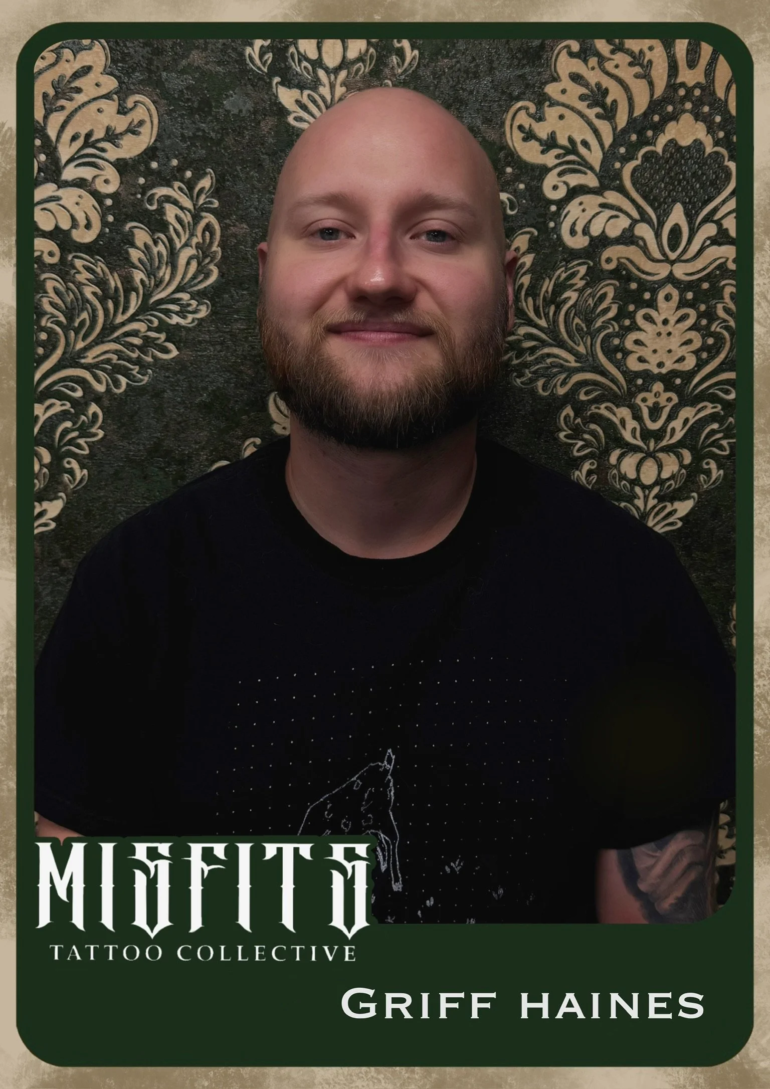 A man with a beard and bald head smiling in front of a decorative floral patterned background, wearing a black T-shirt with a unicorn design and tattoo on the arm, framed as a member of the Moftto Tattoo Collective, with the name Griff Haines on a label.