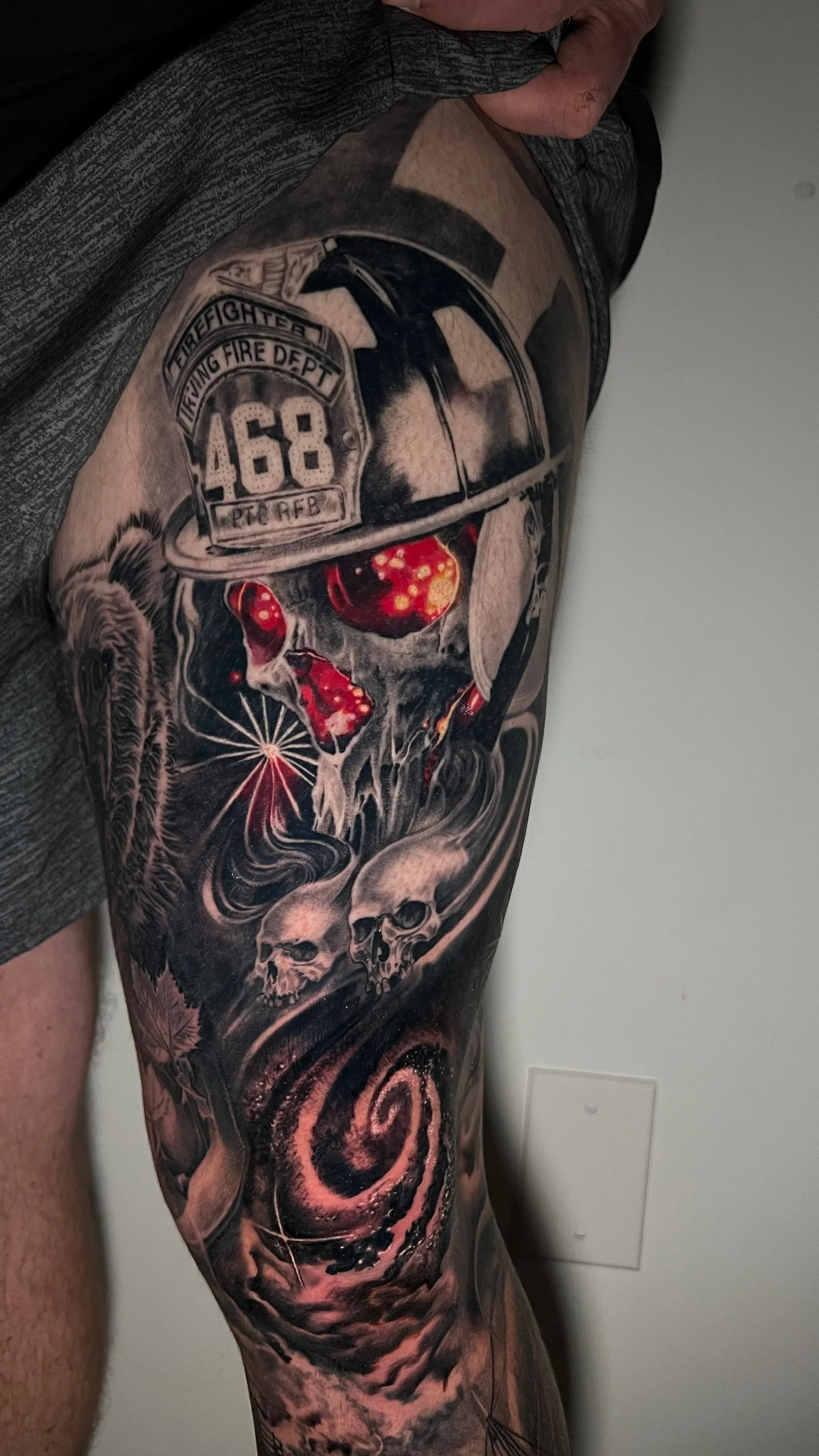 A detailed black and gray tattoo on a person's thigh depicting a firefighter's helmet, a skull with glowing eyes, two smaller skulls, and swirling cosmic patterns.