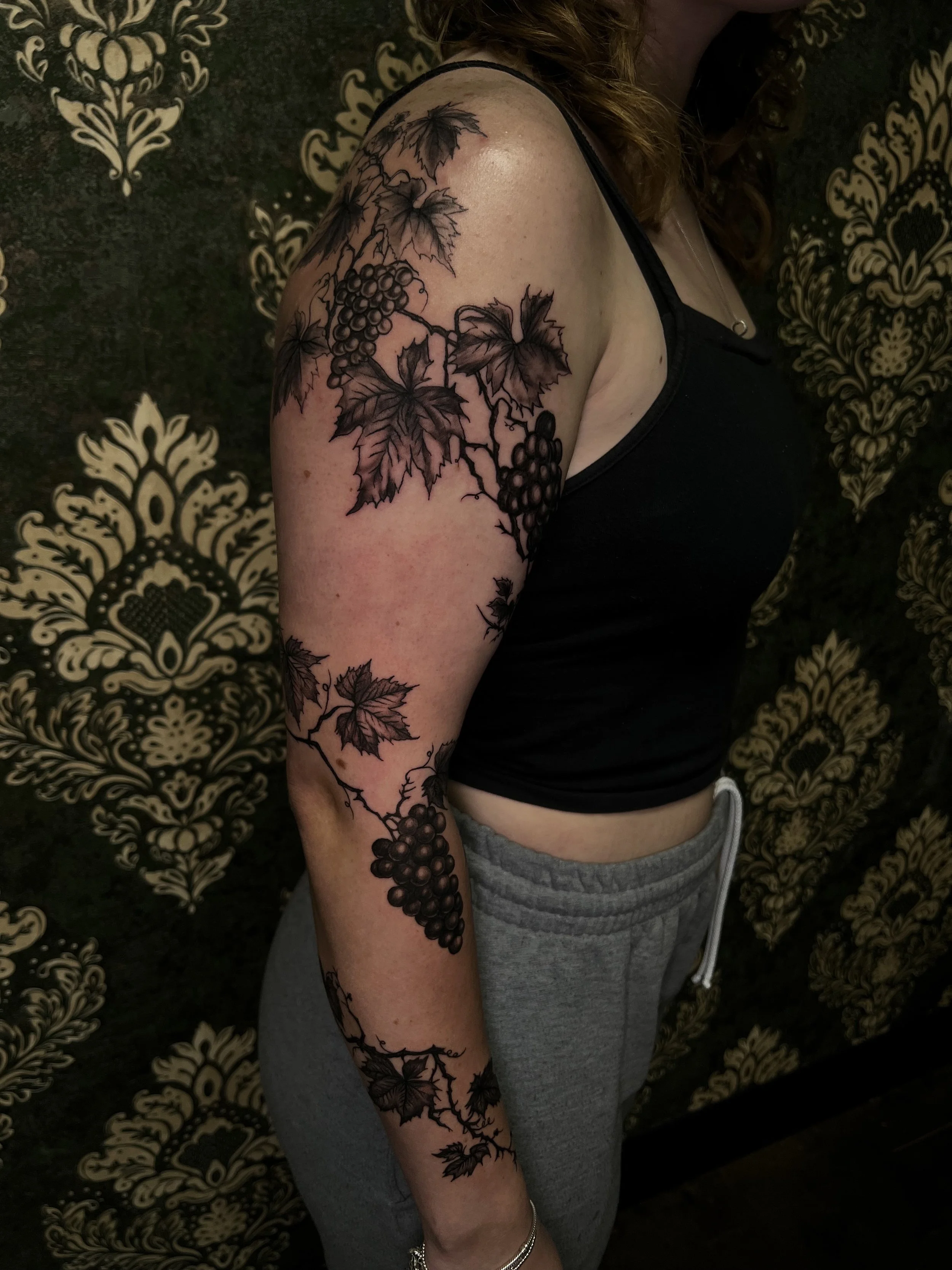 A woman with a large black tattoo of grapevines, leaves, and grapes on her upper arm, shoulder, and forearm, wearing a black top and gray sweatpants, standing against a patterned black and gold wallpaper background.
