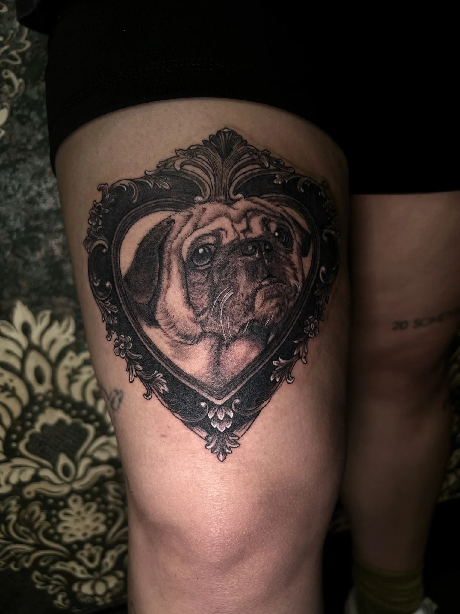 Tattoo of a dog's face inside an ornate heart-shaped frame on a person's thigh.