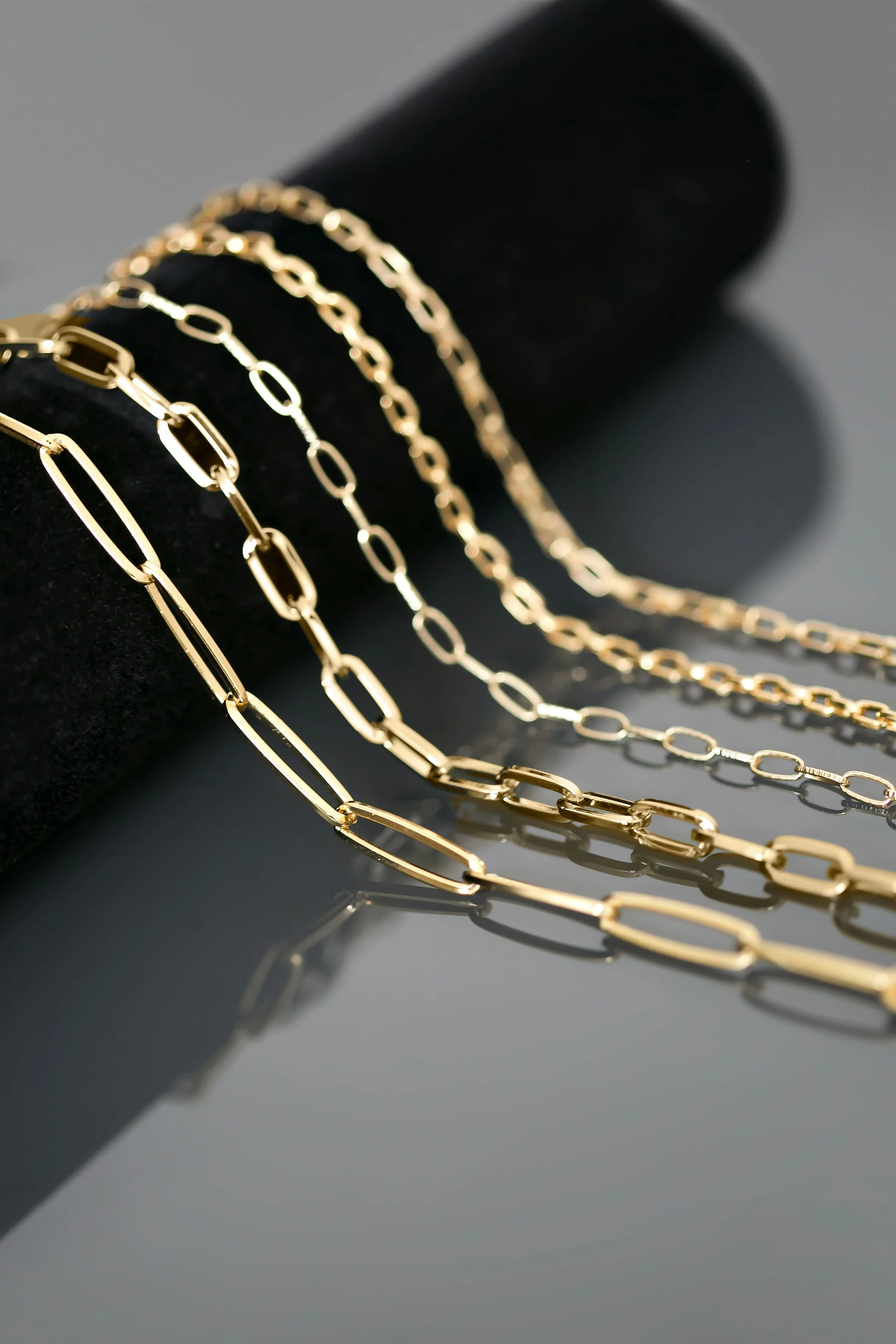 Multiple gold chain necklaces with elongated links displayed on a black velvet bust, with their reflections visible on a glossy surface.