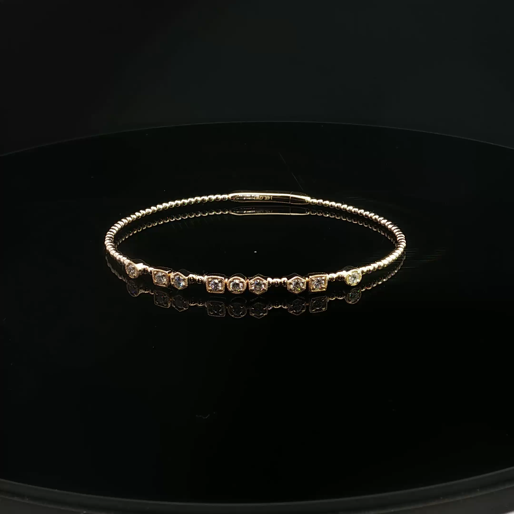 A black surface with a gold bracelet resting on it, with a reflection of the bracelet visible on the surface.