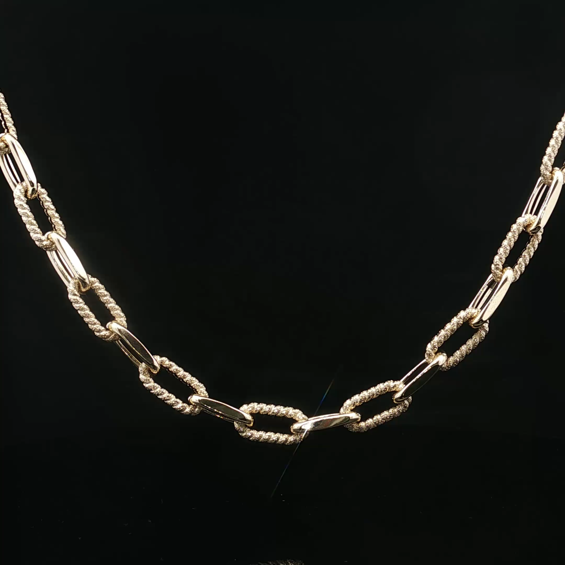 Close-up of a necklace with intertwined gold paperclip links on a black background.