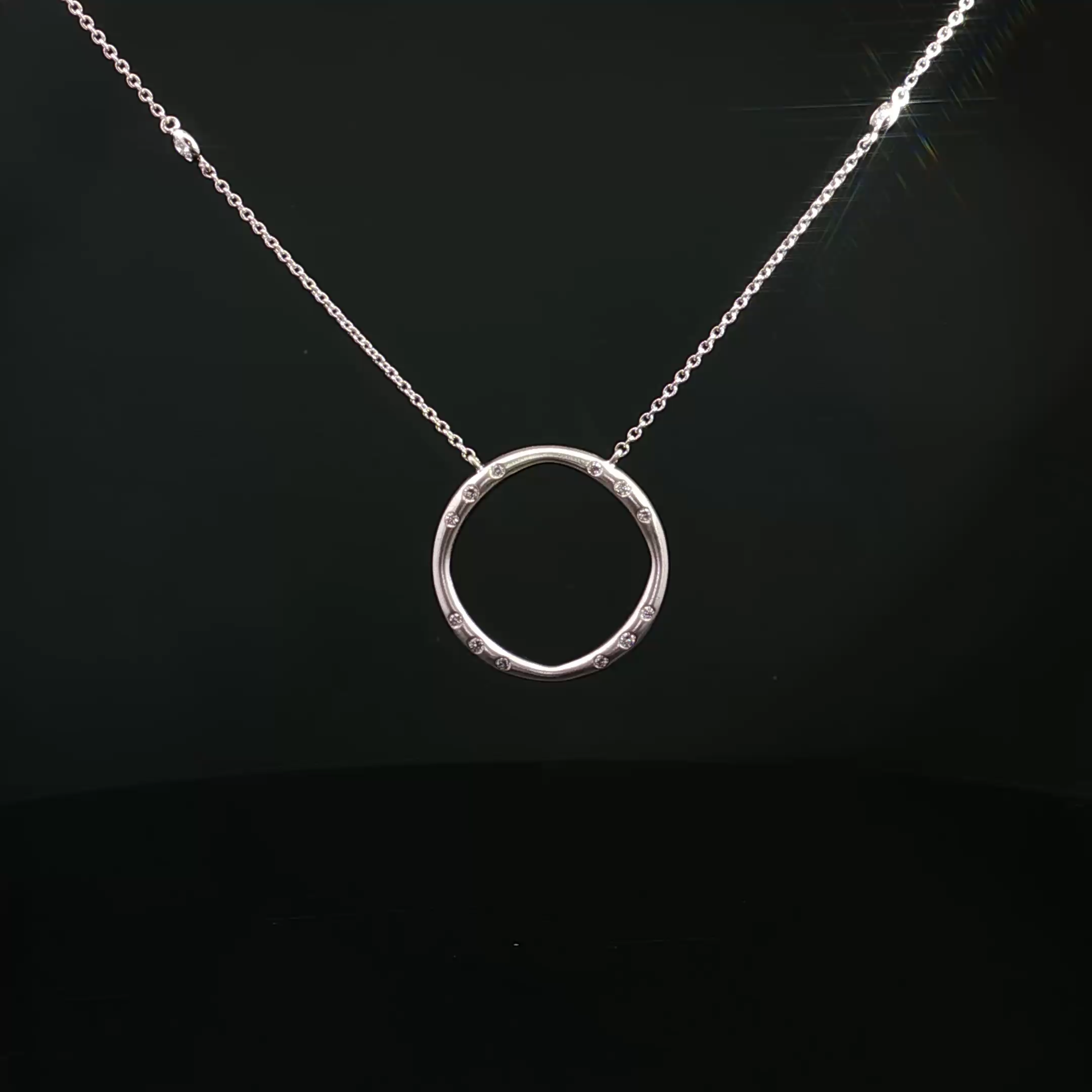 A white gold or silver necklace with a round pendant, decorated with small diamonds, hanging against a black background.
