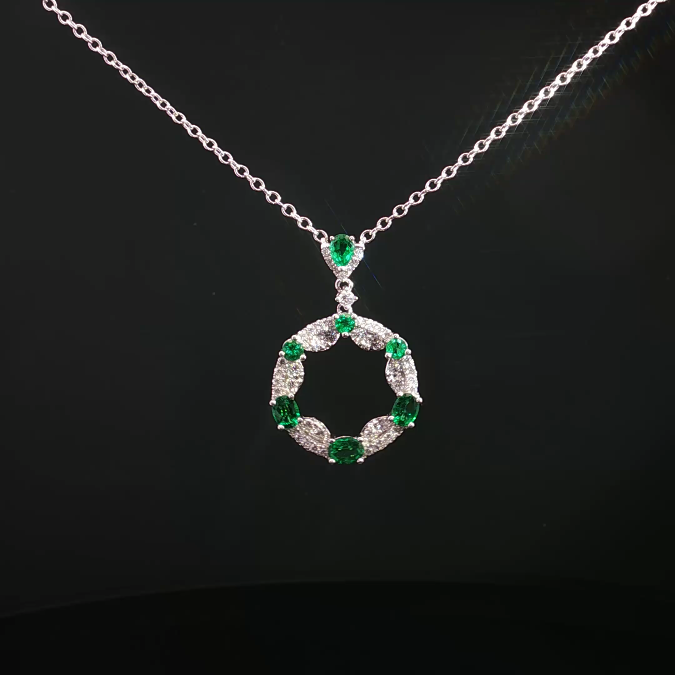 A white gold or silver necklace with green emerald gemstones and diamond accents against a black background.
