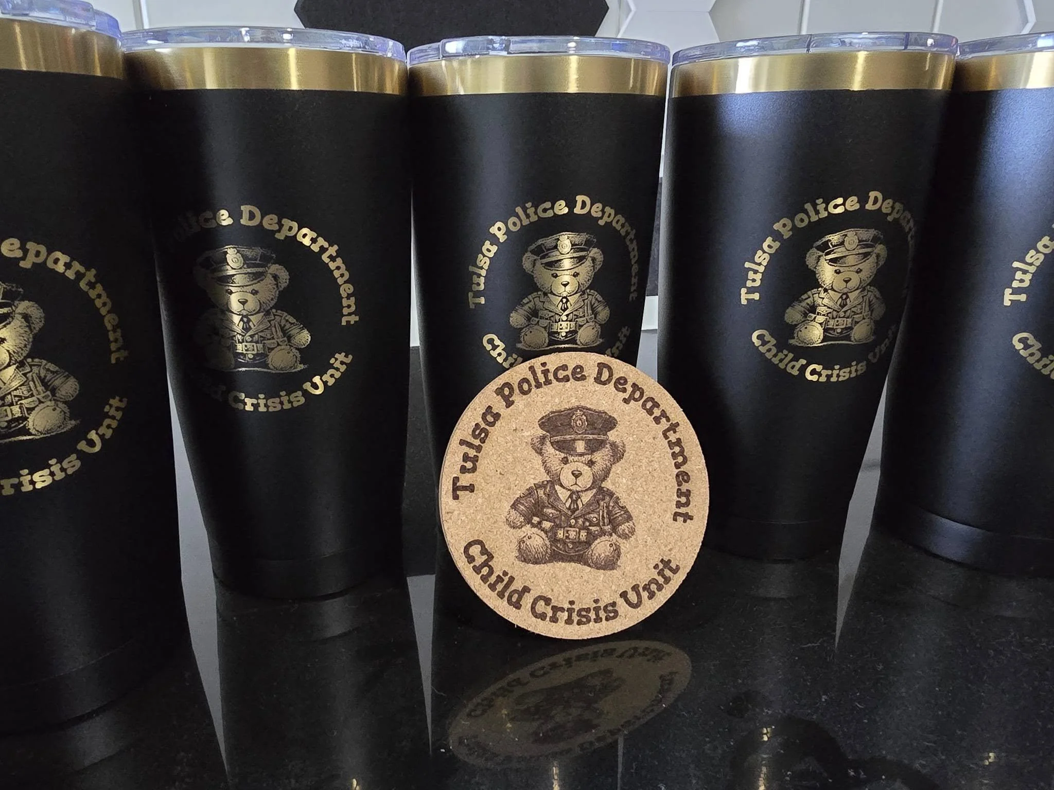 Six black tumblers with a gold logo of a teddy bear in a police uniform, labeled Tulsa Police Department Child Crisis Unit, with one tumbler leaning against others and a circular patch or sticker with the same logo in front.