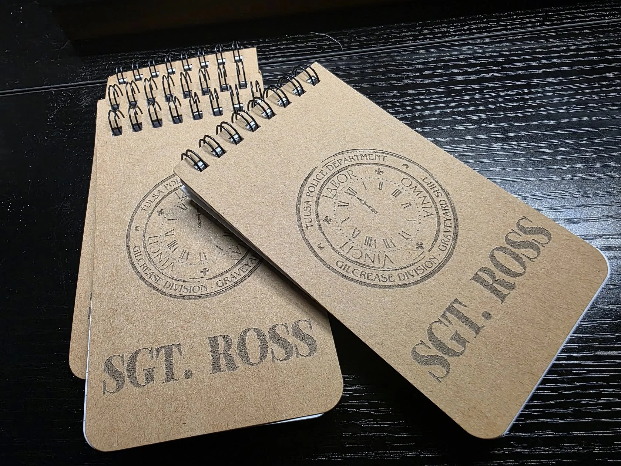 Two spiral notebooks with a police badge stamp and 'SGT. ROSS' printed on the covers, placed on a dark wooden surface.
