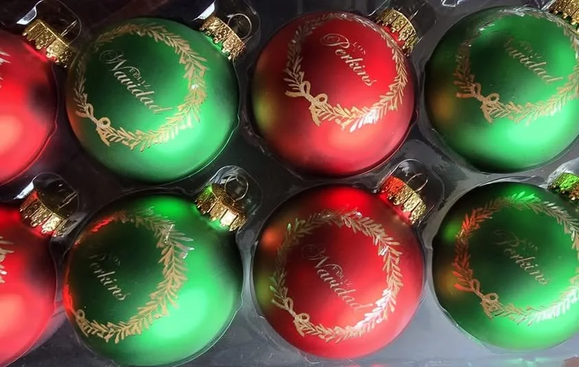 Six Christmas ornaments, three green and three red, with gold writing and decorative wreaths, in a plastic container.