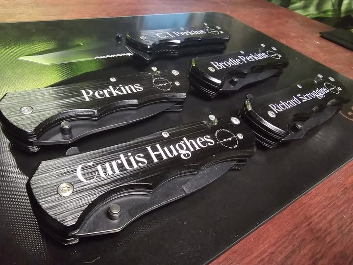 Collection of black multi-tool pocket knives with engraved names, some with visible blades, on a black surface.