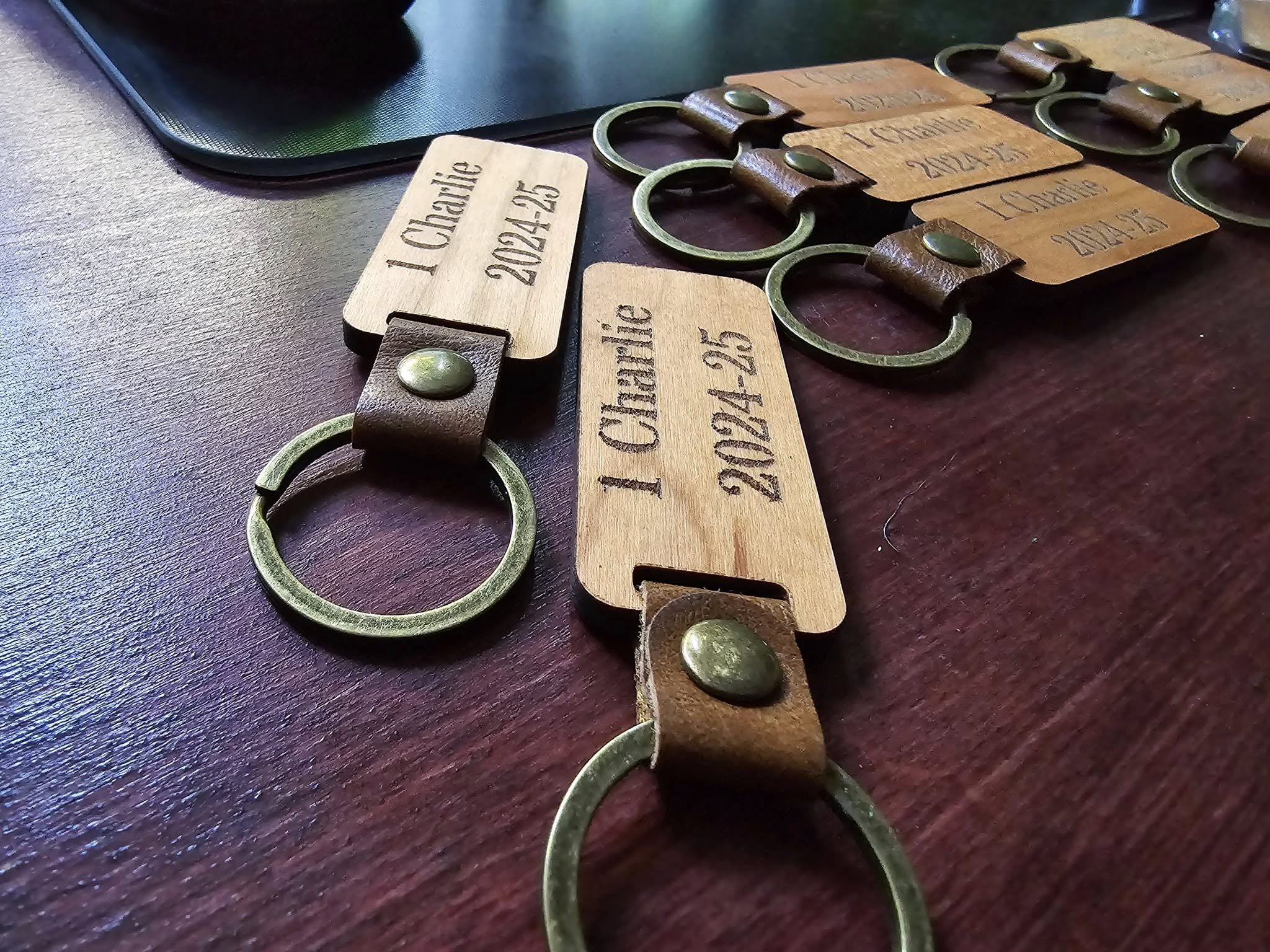 Multiple keychains with wood and leather tags engraved with names and dates, laid out on a dark wooden table.