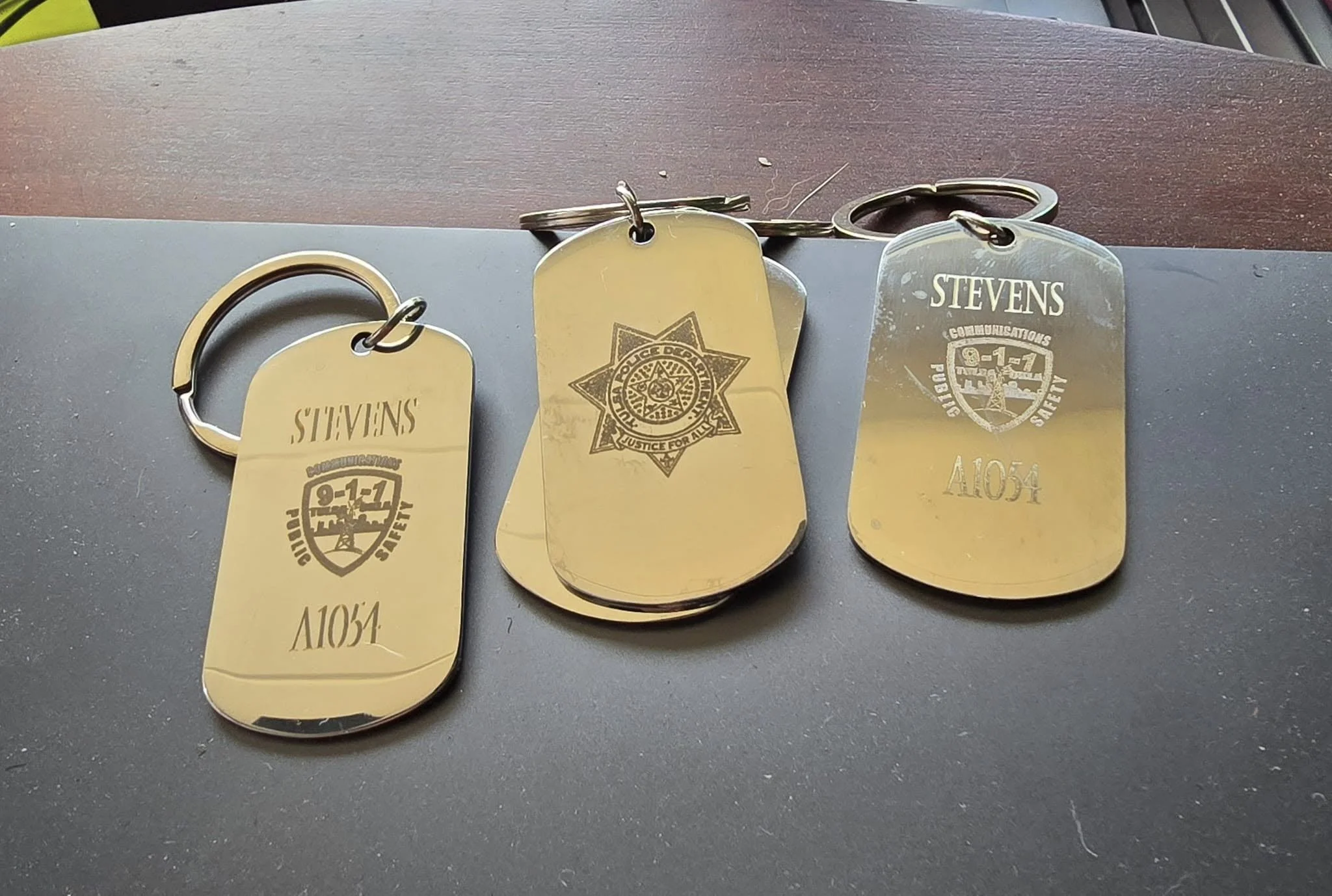Three metal tags with engraved markings, attached to key rings, placed on a dark surface with a wooden background.