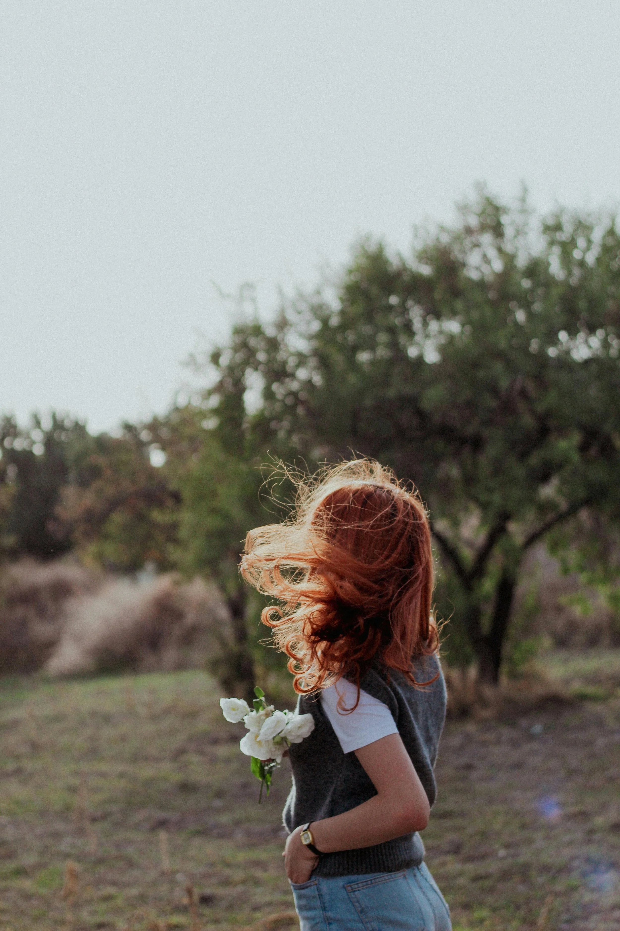 A woman with red hair standing outdoors in a natural setting, holding white flowers behind her back, with trees and an overcast sky in the background.