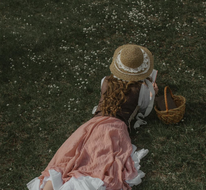 Girl with curly hair, wearing a straw hat, pink skirt, and white top, sitting on grass with white flowers, using a mobile device, with a basket nearby.