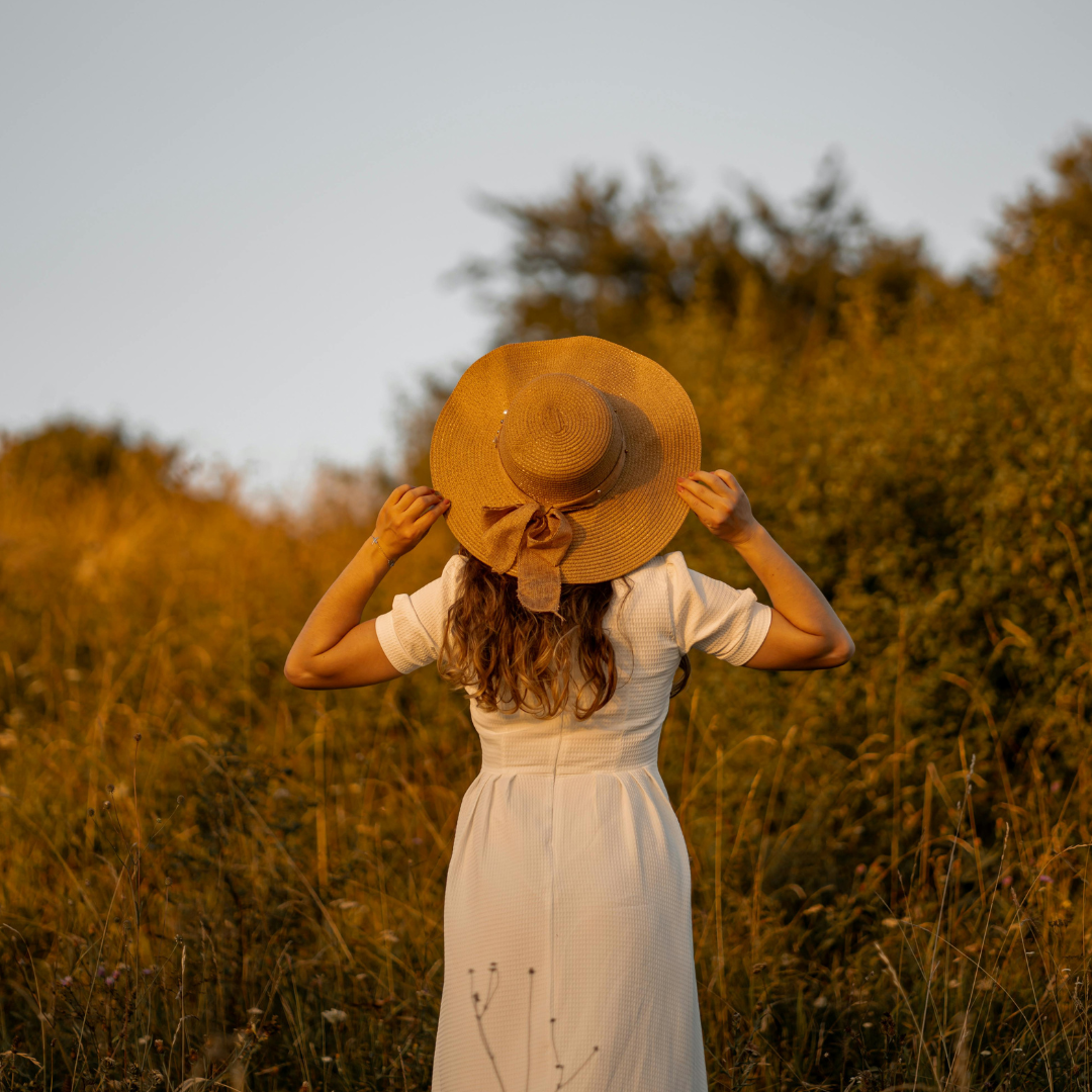 A woman in a white dress holding a wide-brimmed straw hat with a bow, standing in a field during sunset with trees in the background.