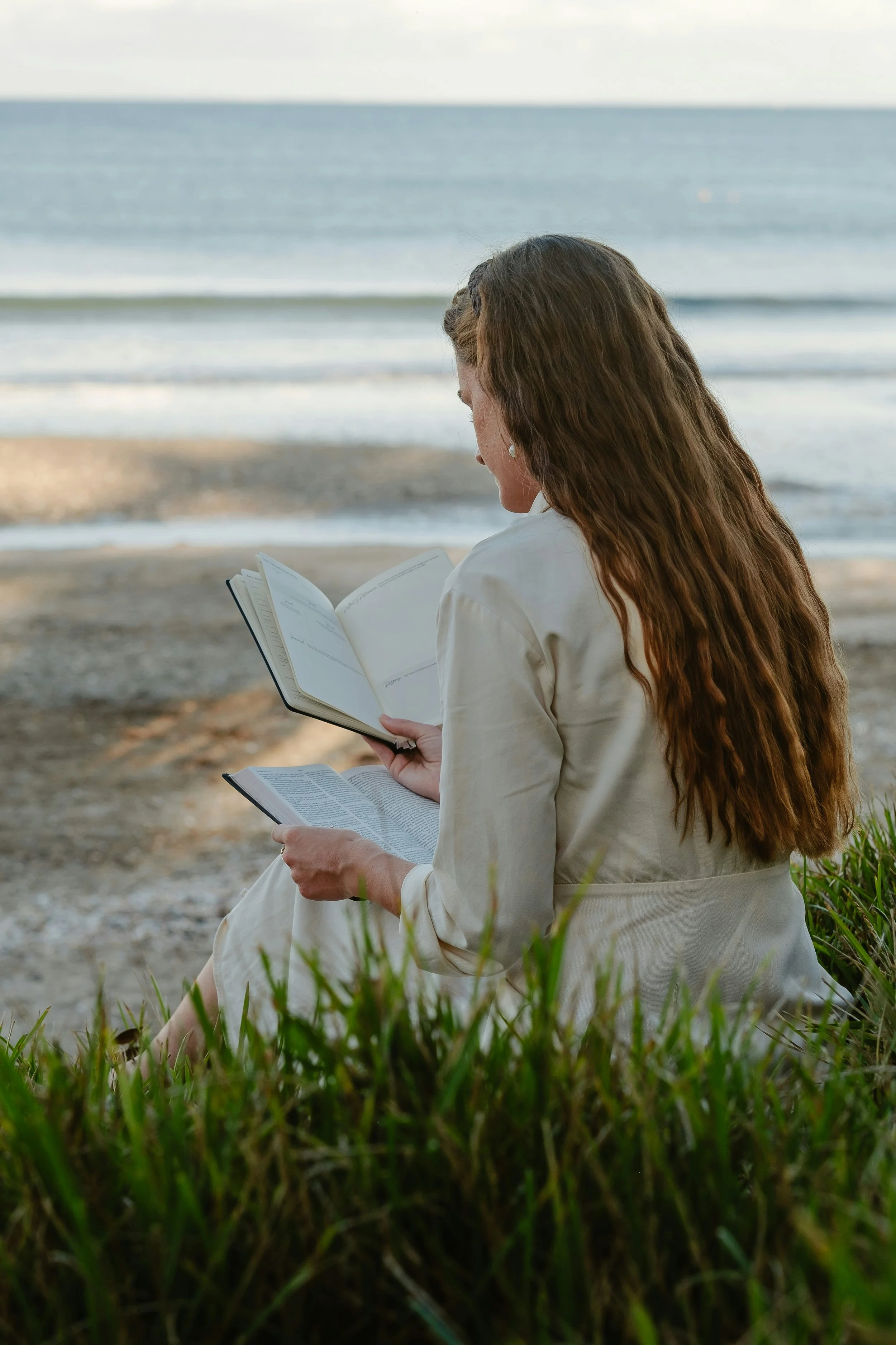 A woman with long, wavy reddish-brown hair sitting on grass near a beach, reading a book against the backdrop of ocean waves.