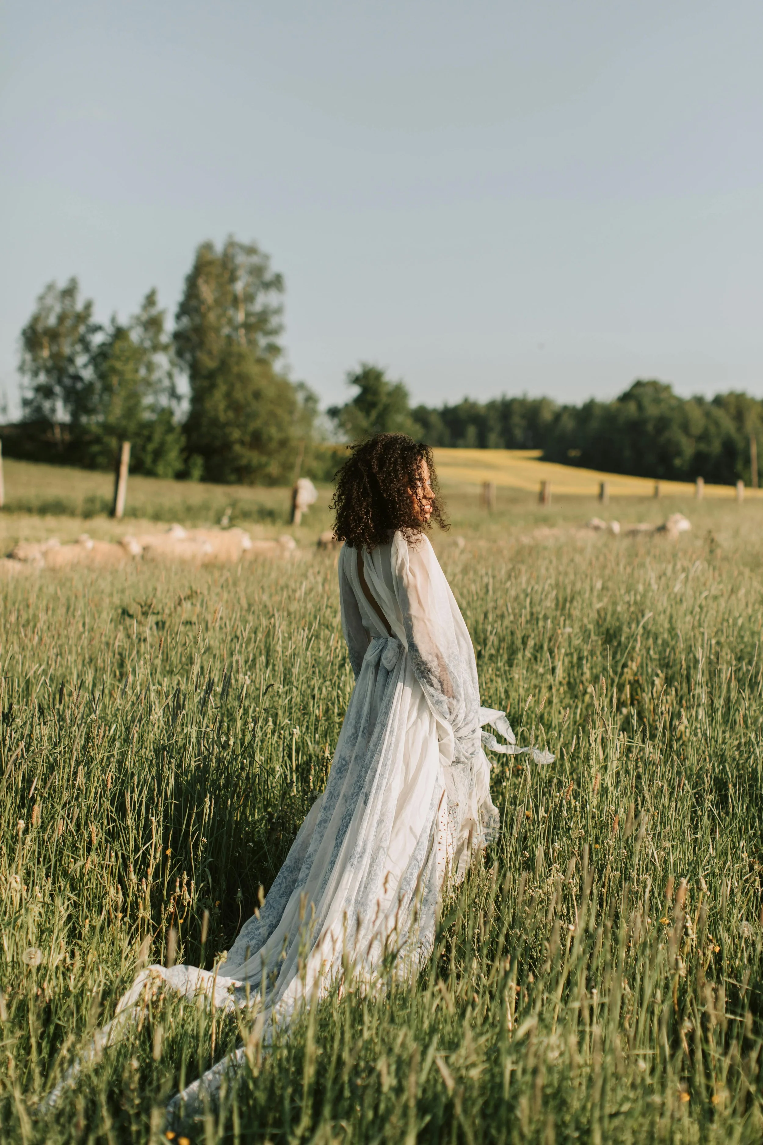 Woman with curly hair in a white dress standing in a grassy field with trees and a blue sky in the background.