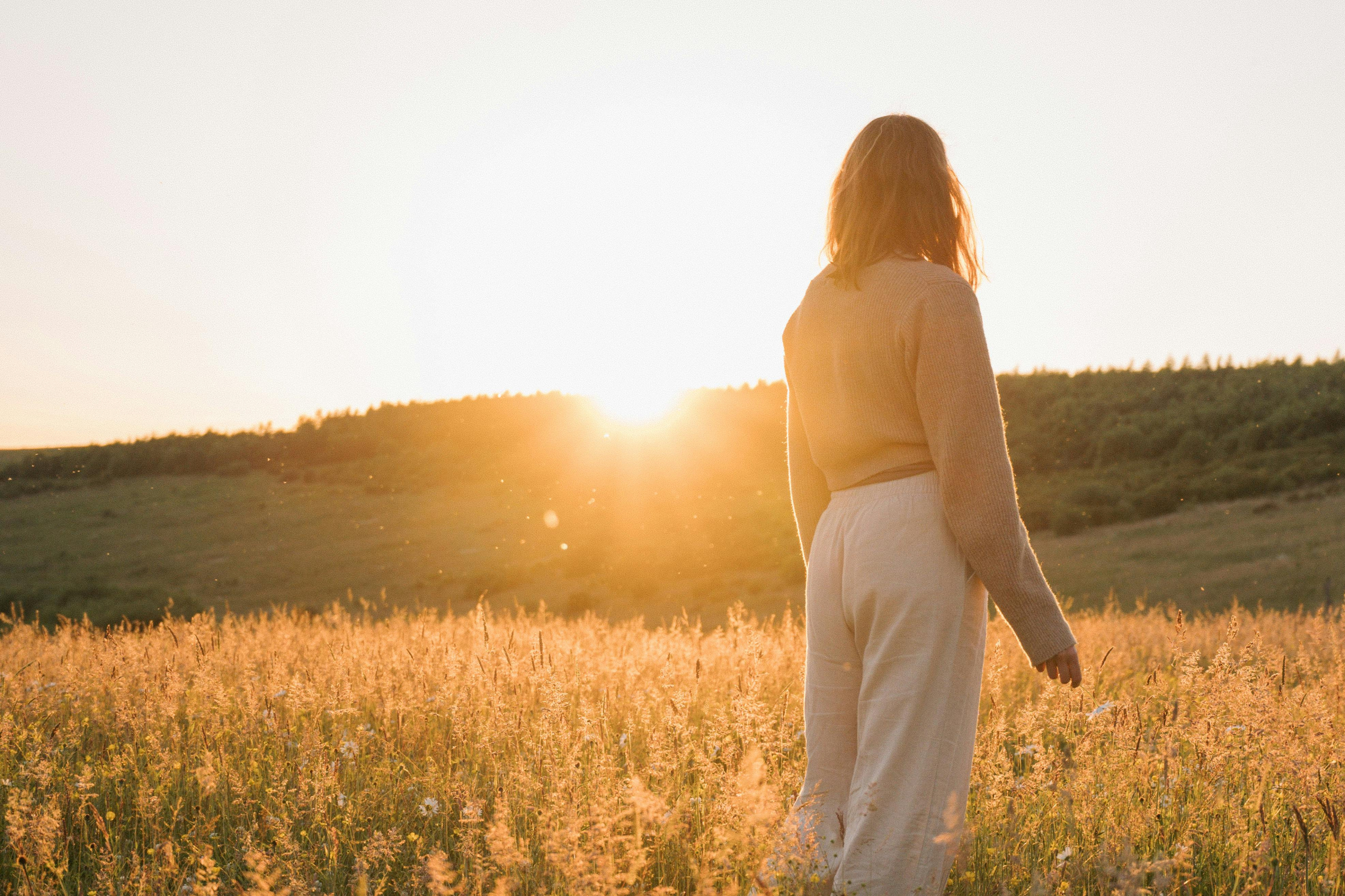 A woman standing in a field of tall grass during sunset with her back to the camera.
