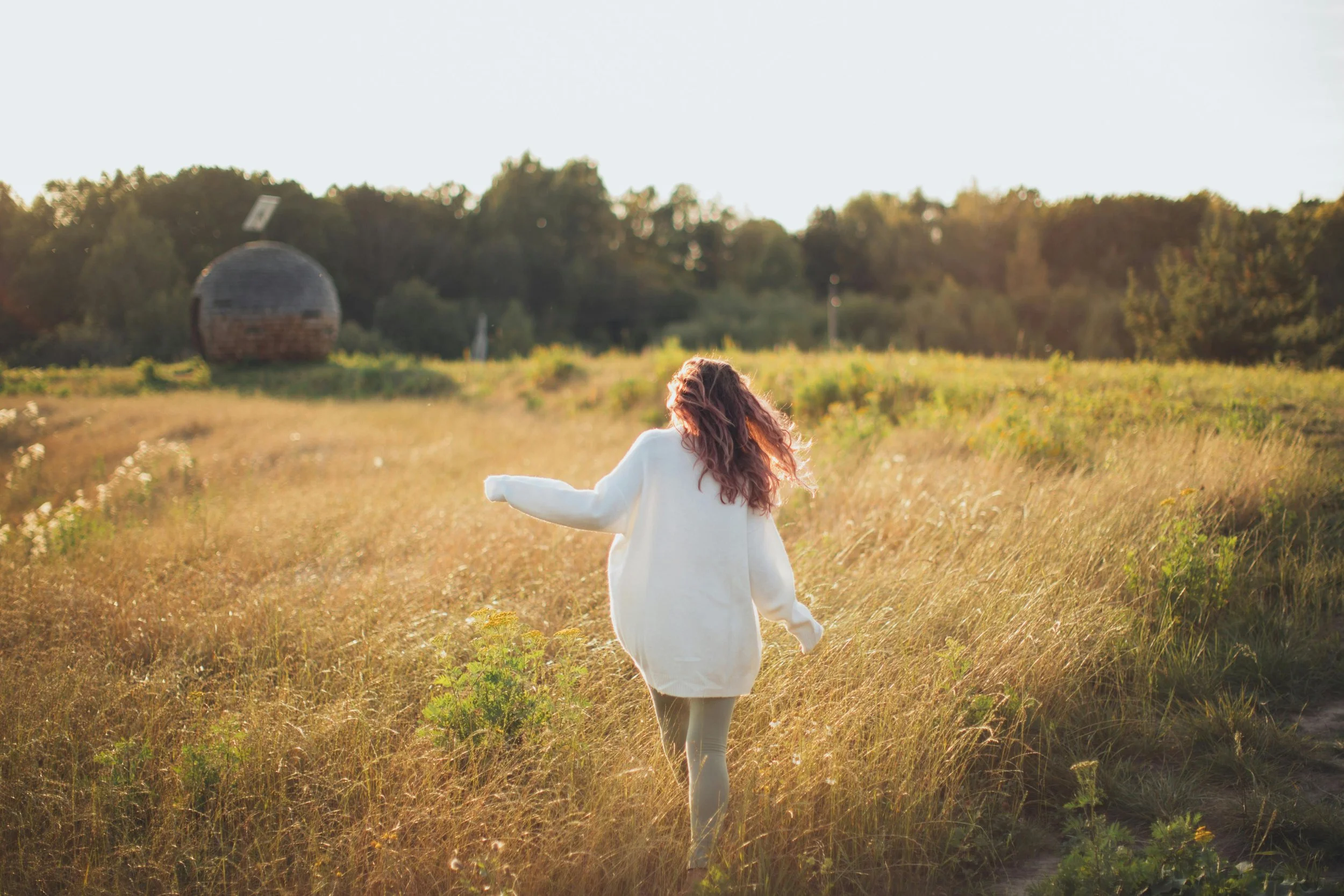 A woman with long, wavy hair walking through a grassy field during sunset, wearing a white oversized sweater.