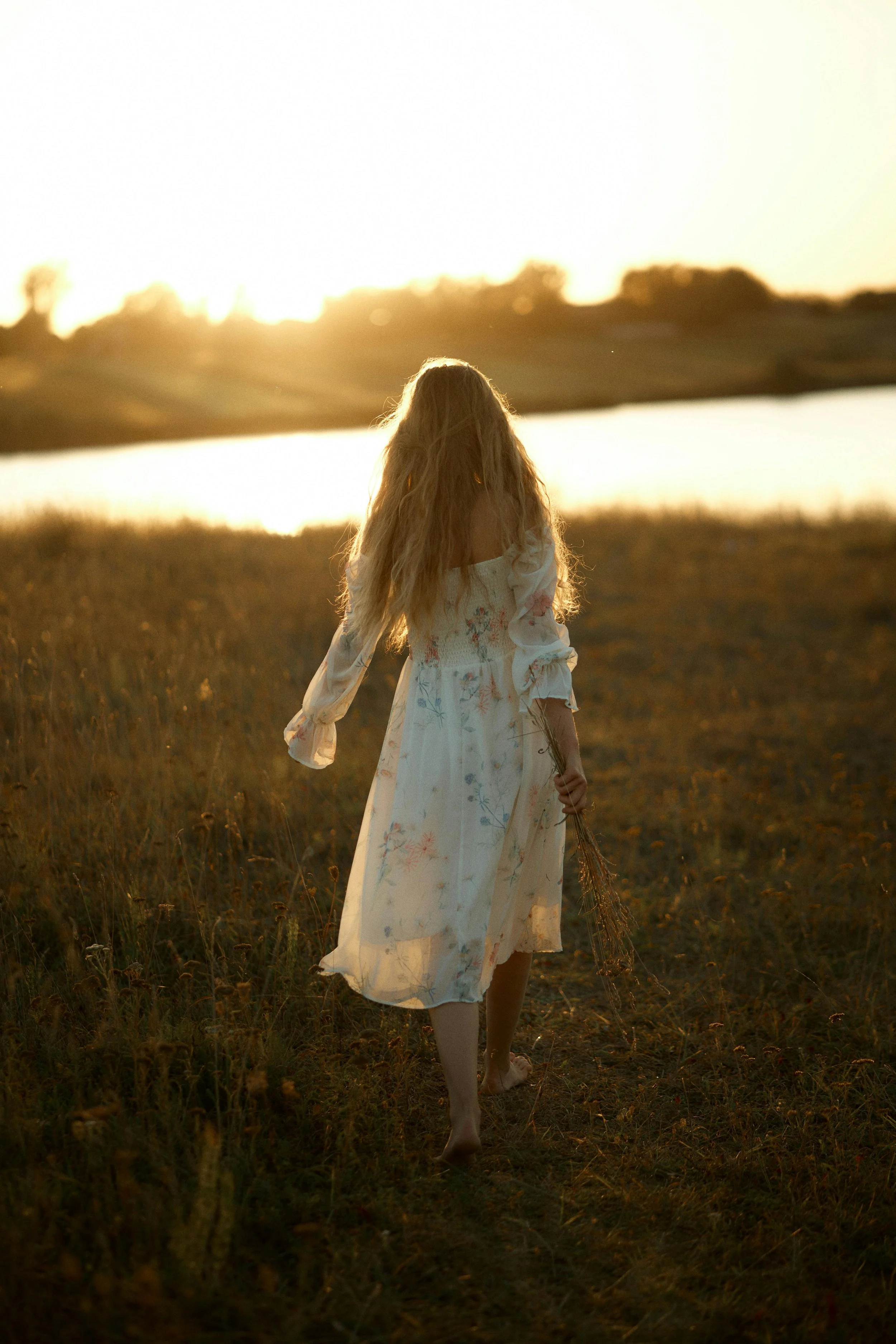 A girl in a white, floral dress walking on a grassy path near a pond during sunset, holding a small bundle of dried plants.