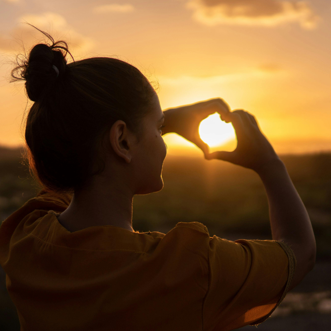 A woman making a heart shape with her hands around the setting sun during sunset.