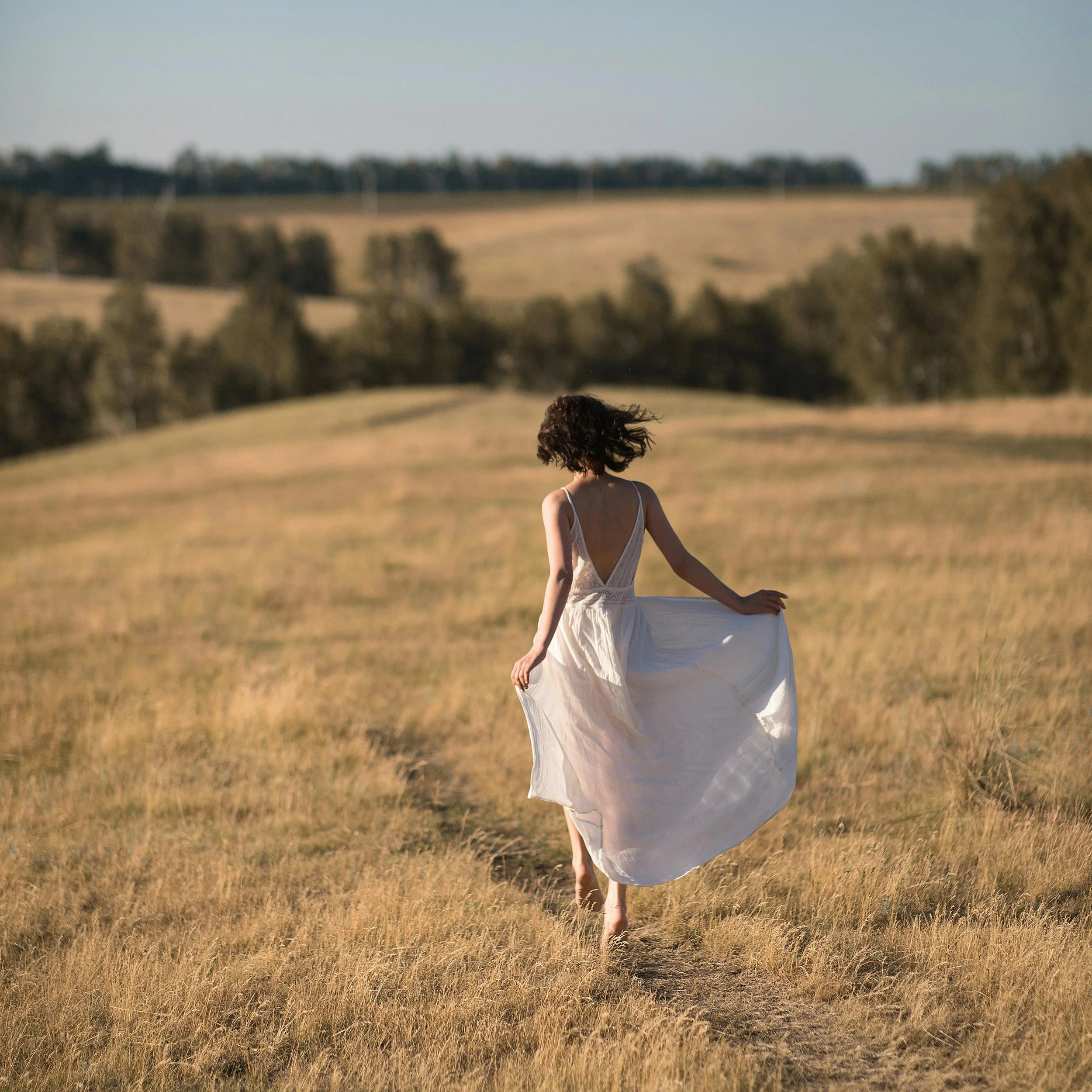 A woman in a white dress walking away through a grassy field with trees in the background.