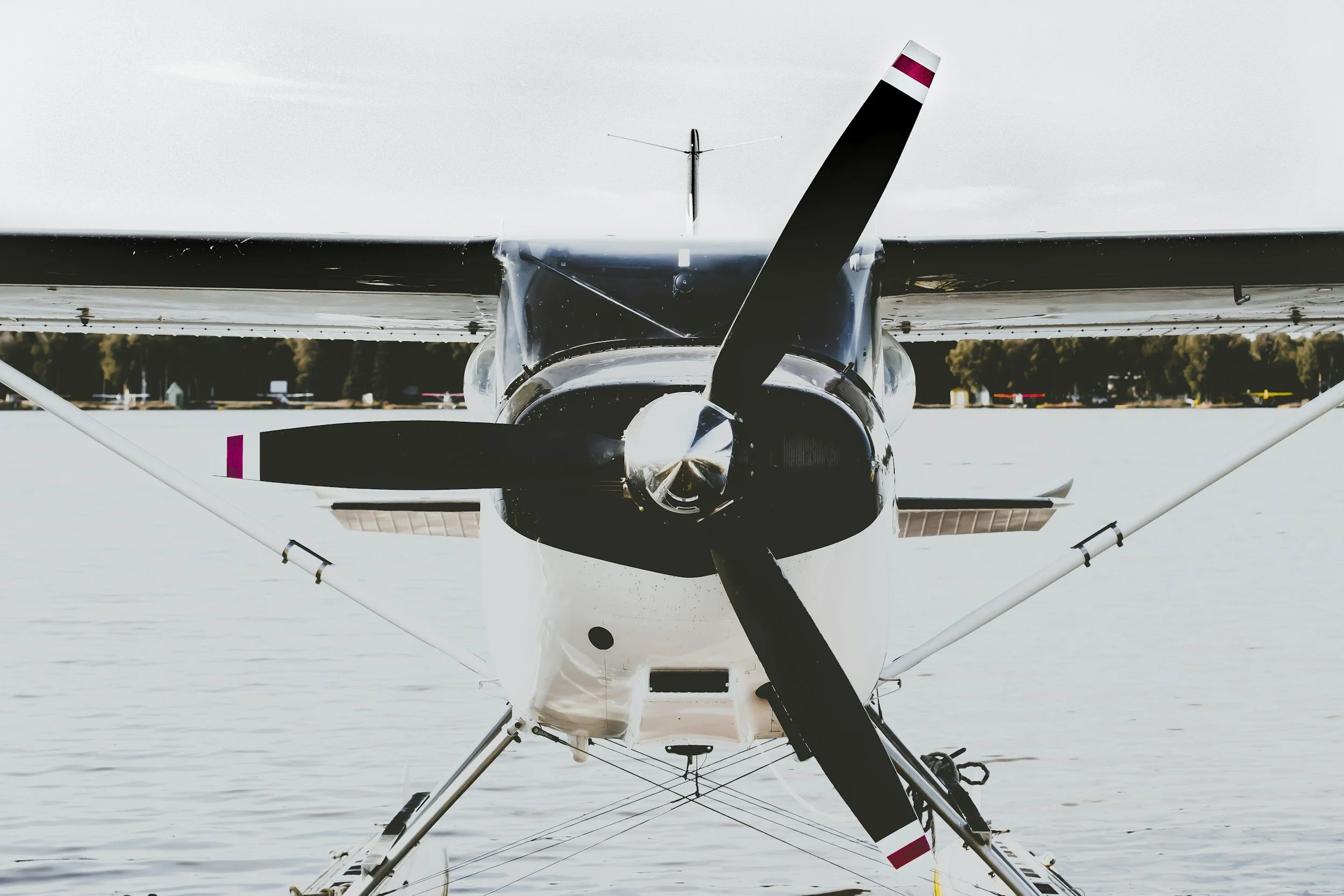 Front view of a seaplane with a black and white body and a three-blade propeller, floating on water with trees and airport structures in the background.