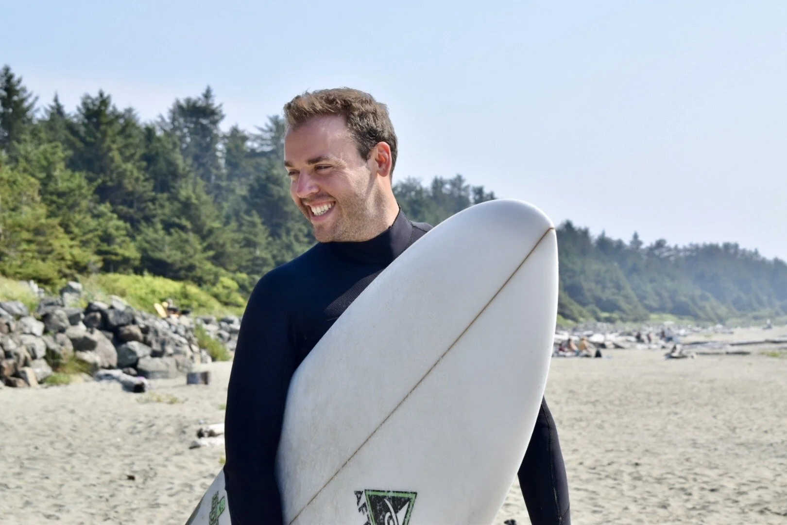 A man in a wetsuit holding a white surfboard on a beach near the ocean with green trees in the background.