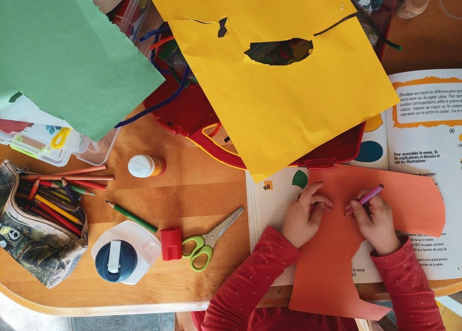 A table covered in craft materials and a child's hands drawing on paper.