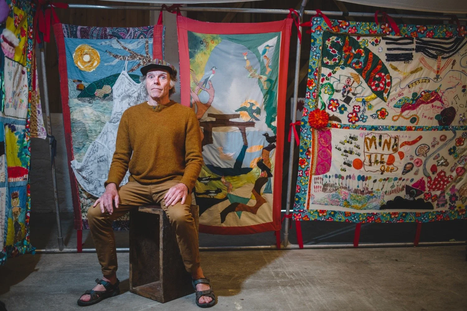 A man sits in front of colourful fabric panels depicting Cornish stories inside Story Shelter.