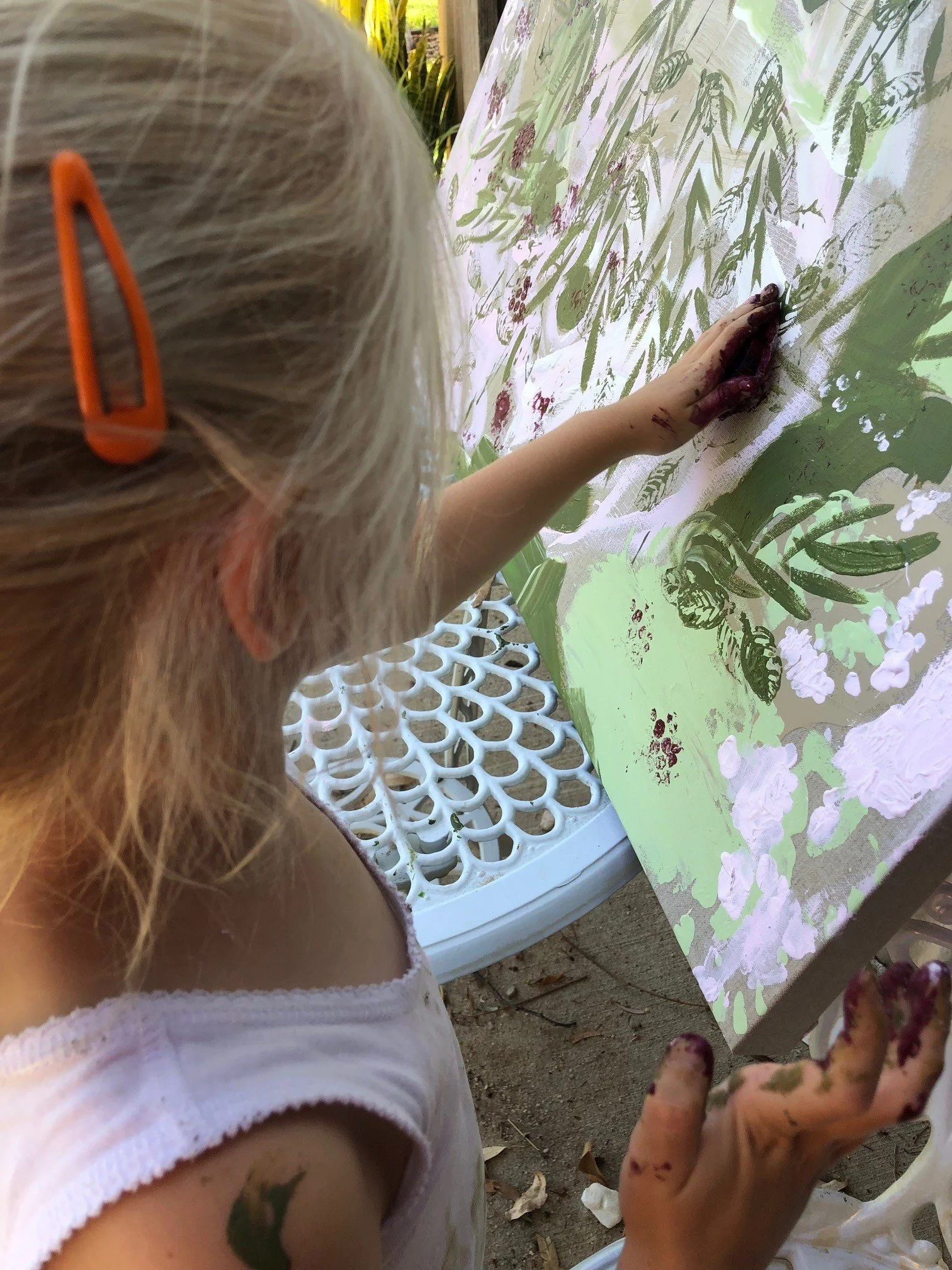 Young girl with blonde hair and an orange hair clip painting on a canvas with green and white colors, her hands covered in paint.