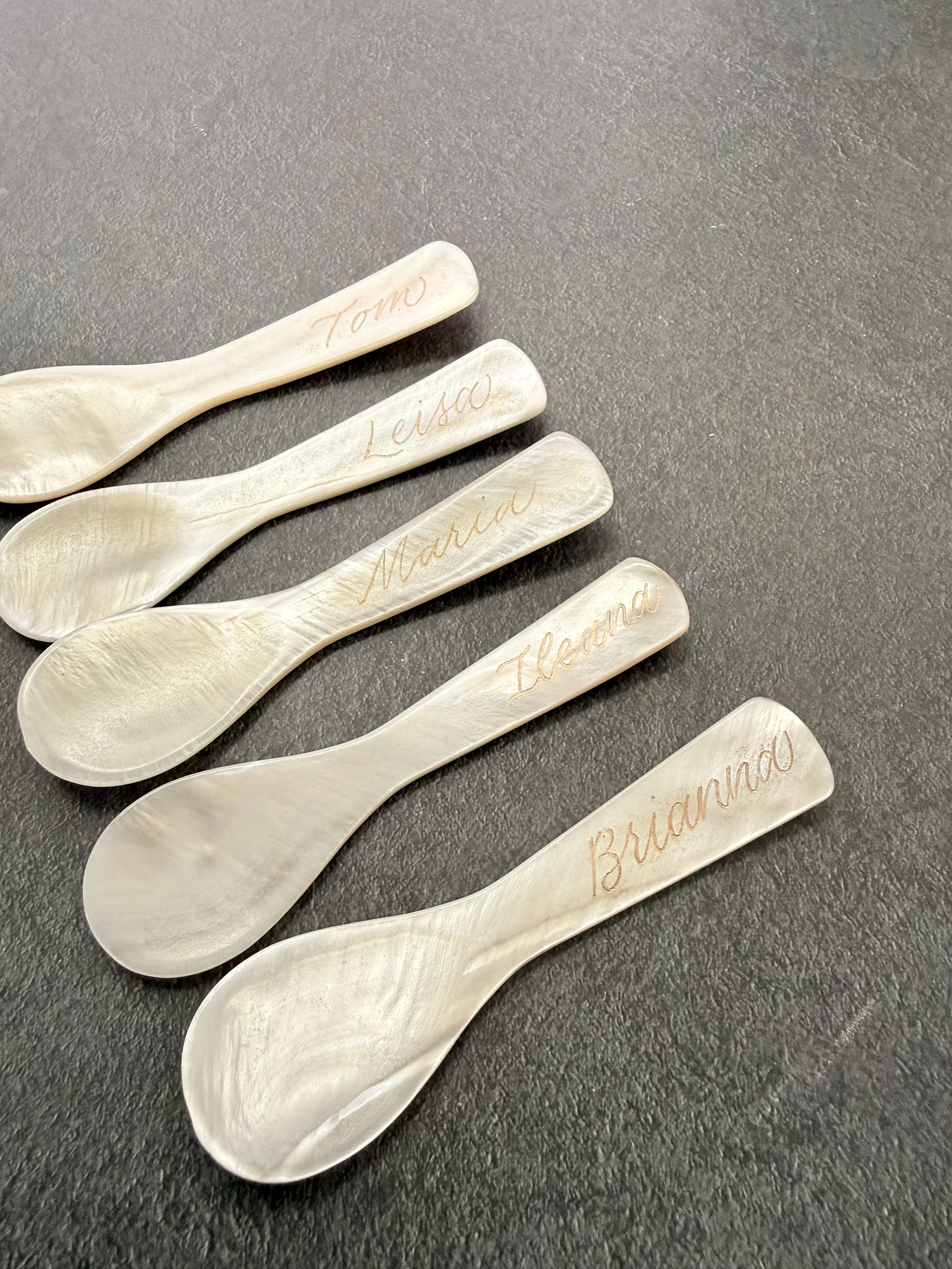Six small wooden spoons on a dark surface, each engraved with a name: Tomo, Lenso, Marid, Island, Ileana, and Briano.