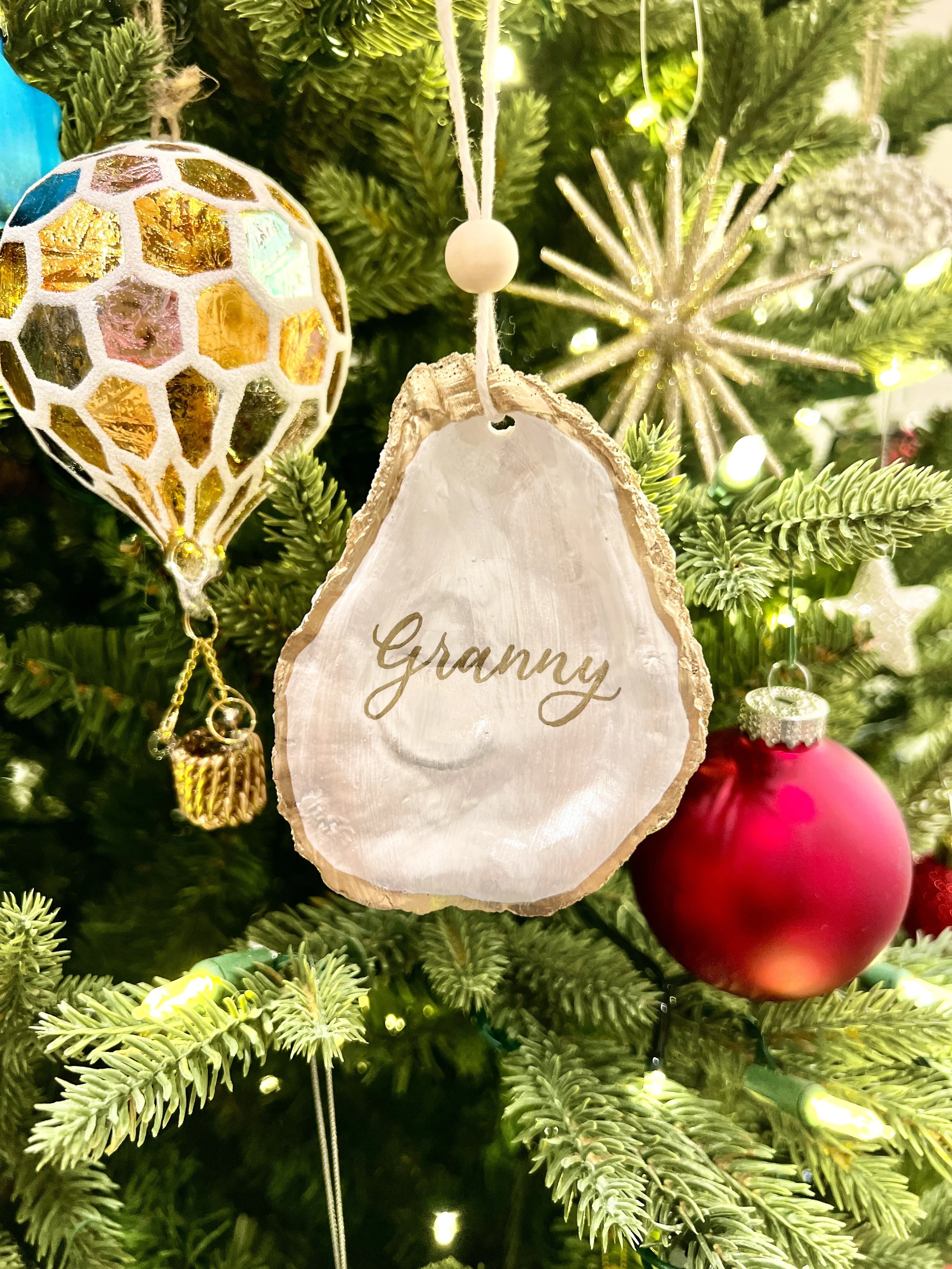 A decorated Christmas tree with a white-marble ornament inscribed with the word 'Granny,' a red bauble, a golden and silver ornament, and a star-shaped ornament, all surrounded by green pine branches and string lights.