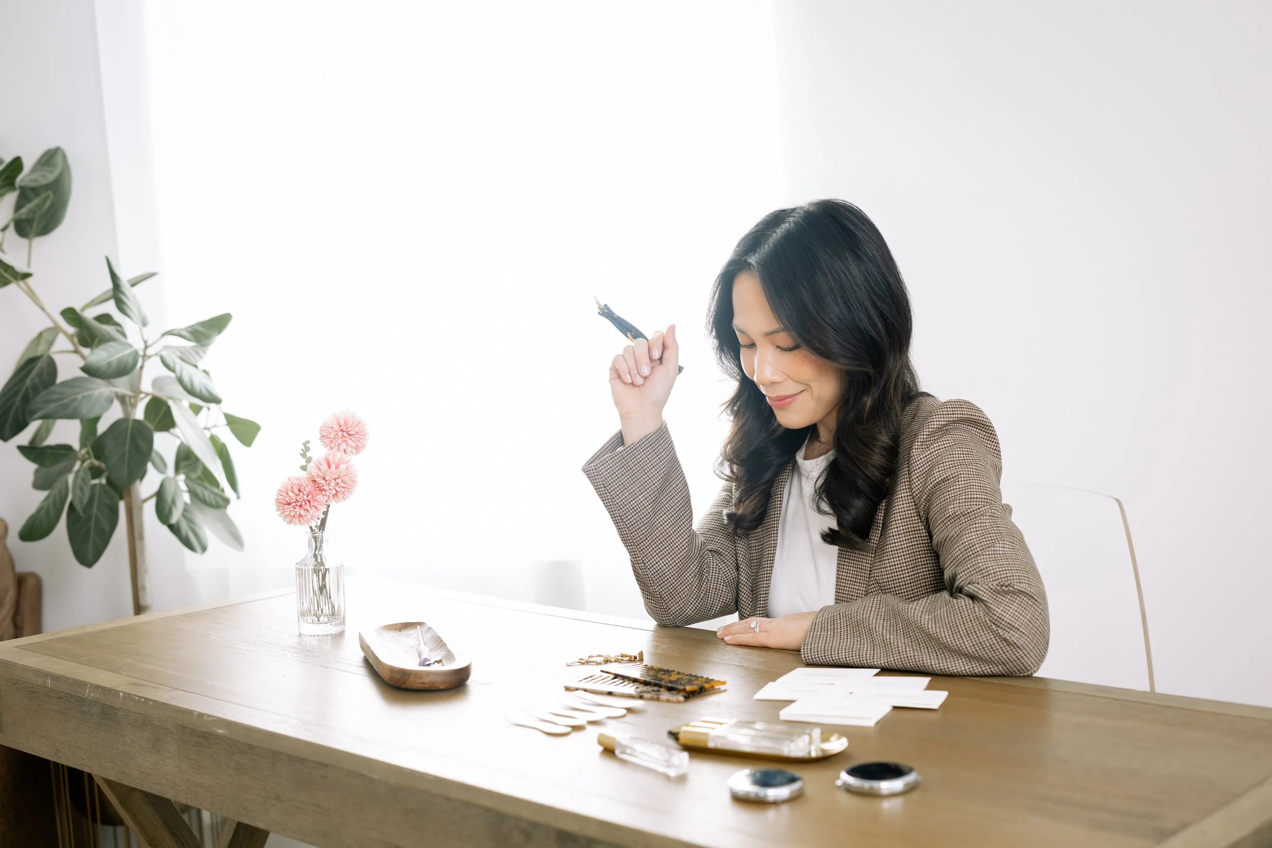 Woman with dark hair in a checked blazer sitting at a wooden table, holding an eyeliner pencil, with makeup products and cards on the table, in a bright room with white walls and a large window.