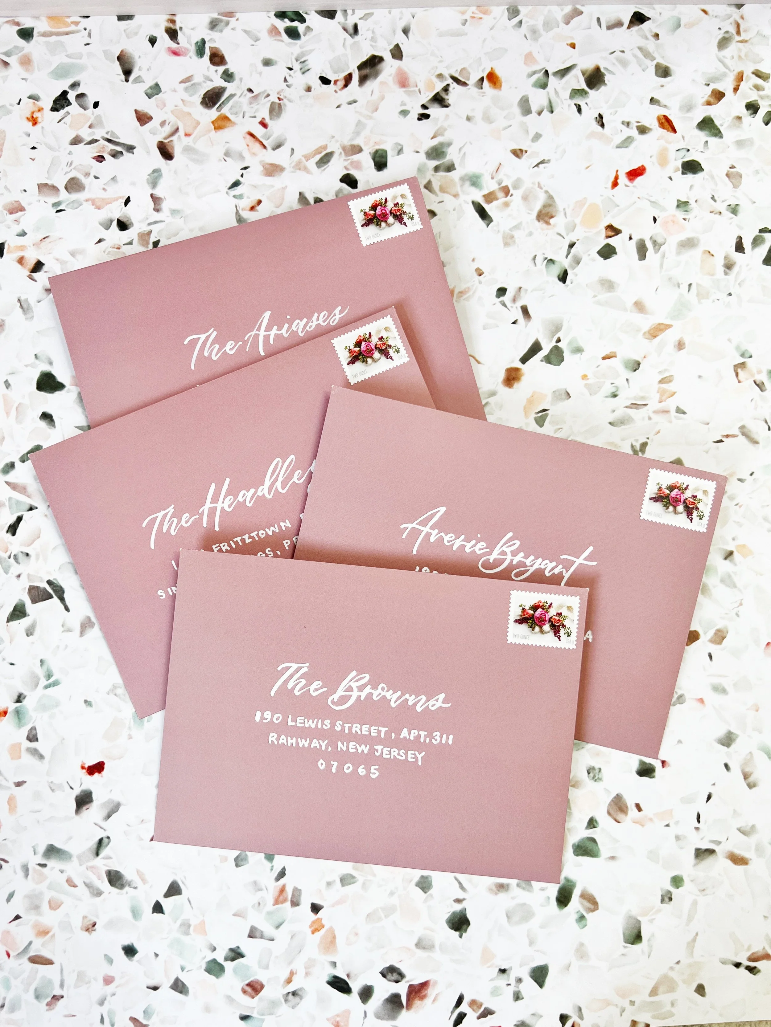 Four pink envelopes with white handwritten-style text and floral stamps on a terrazzo surface. The envelopes are addressed to different recipients, with one showing an address in New Jersey.