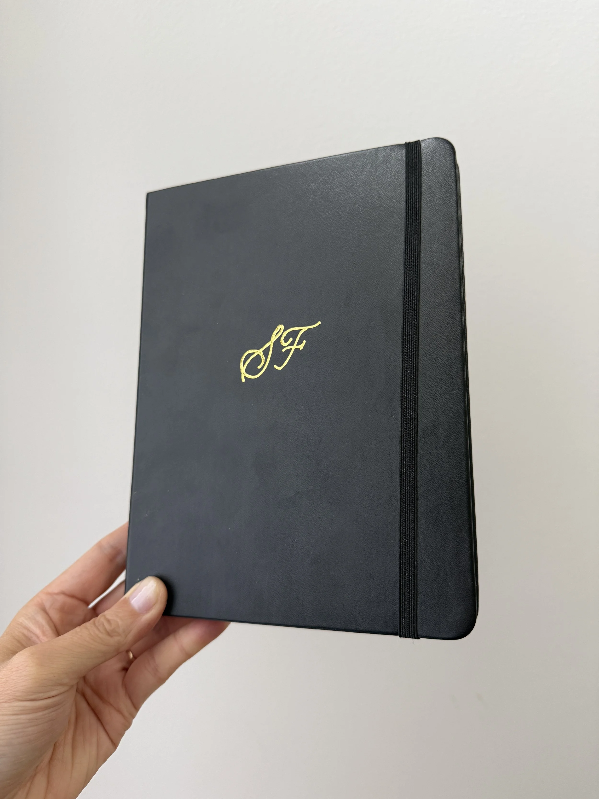 Hand holding a black notebook with a gold monogram 'J F' on the cover.