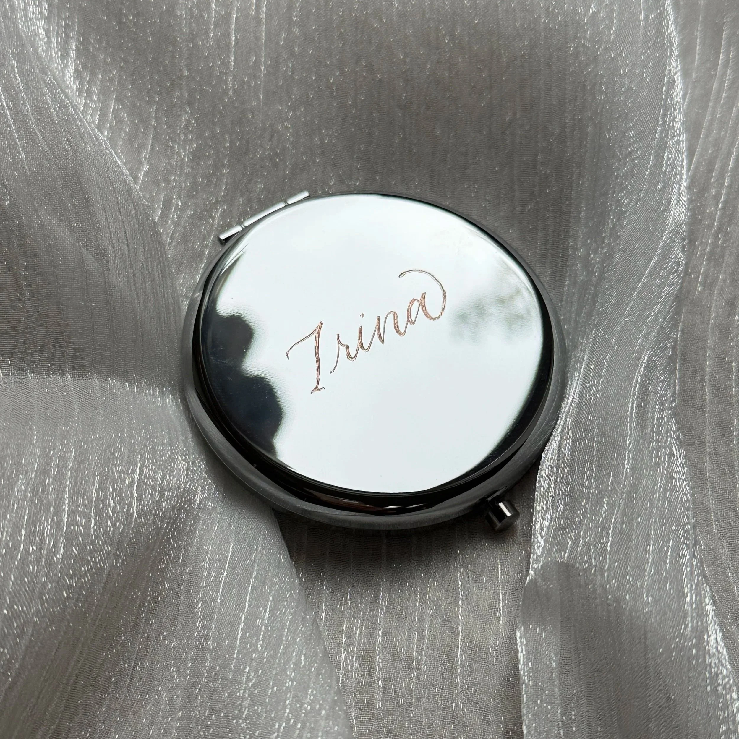 A silver, round compact mirror with the word 'Irina' handwritten on it in cursive, placed on a silver, shimmery fabric.