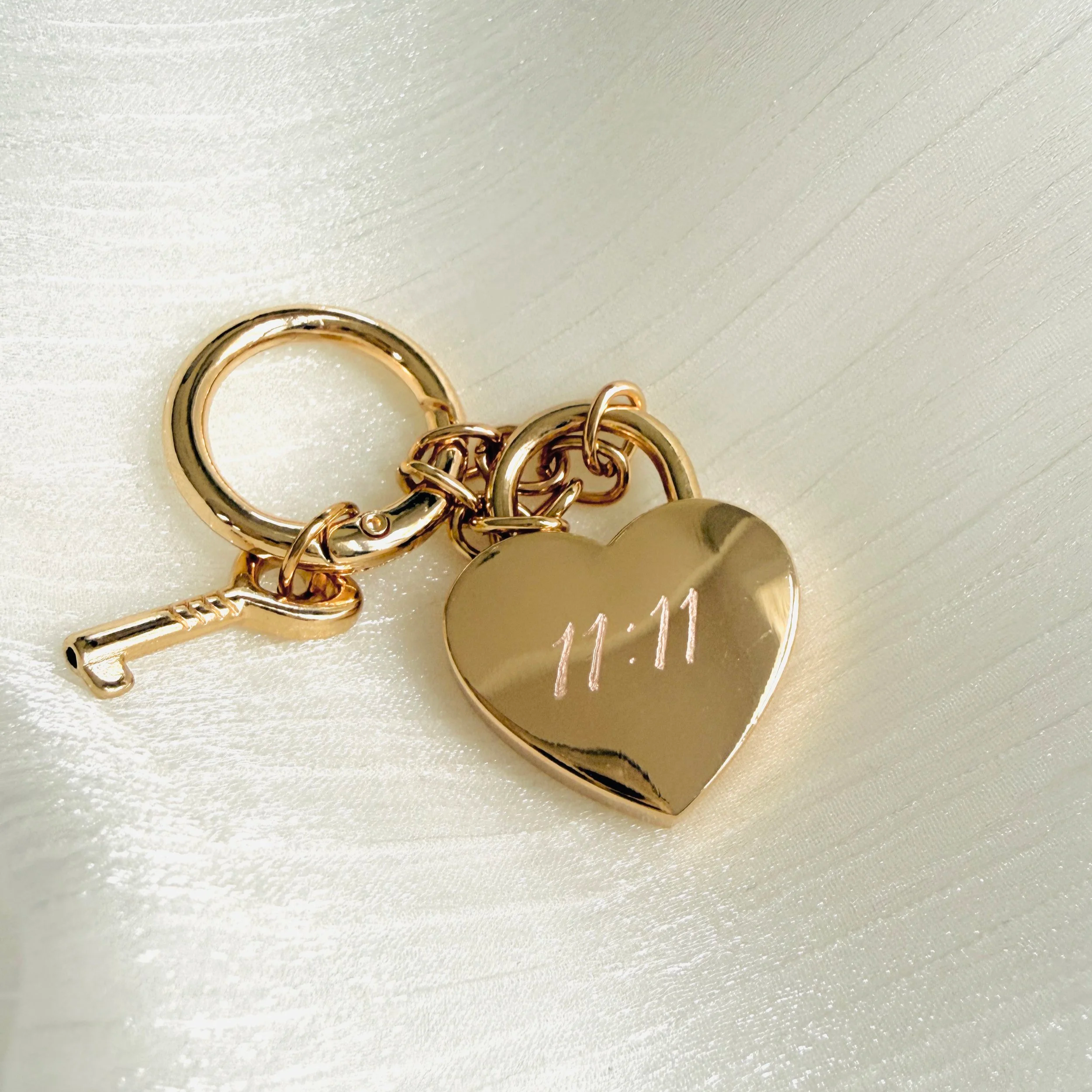A gold keychain with a heart-shaped charm engraved with the time 11:11, a small key charm, and a circular keyring, all placed on a satin fabric surface.